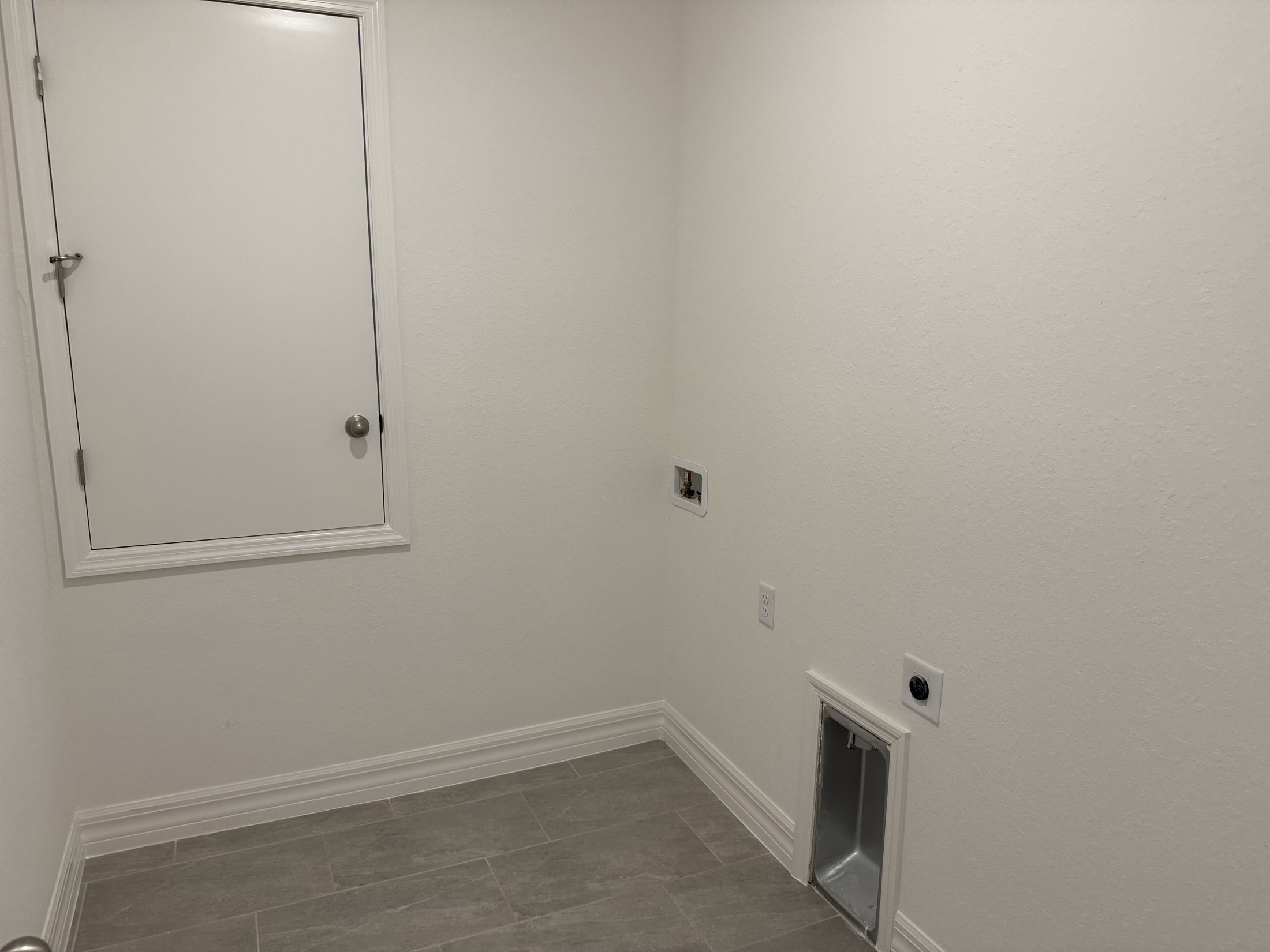 Empty laundry room with white walls, gray tile flooring, and utility hookups.