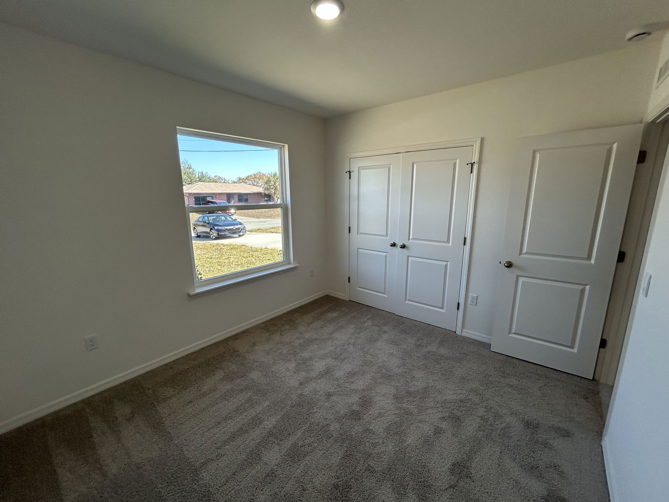 Spacious and bright bedroom featuring a large window with a view, plush carpet flooring, and modern white closet doors.