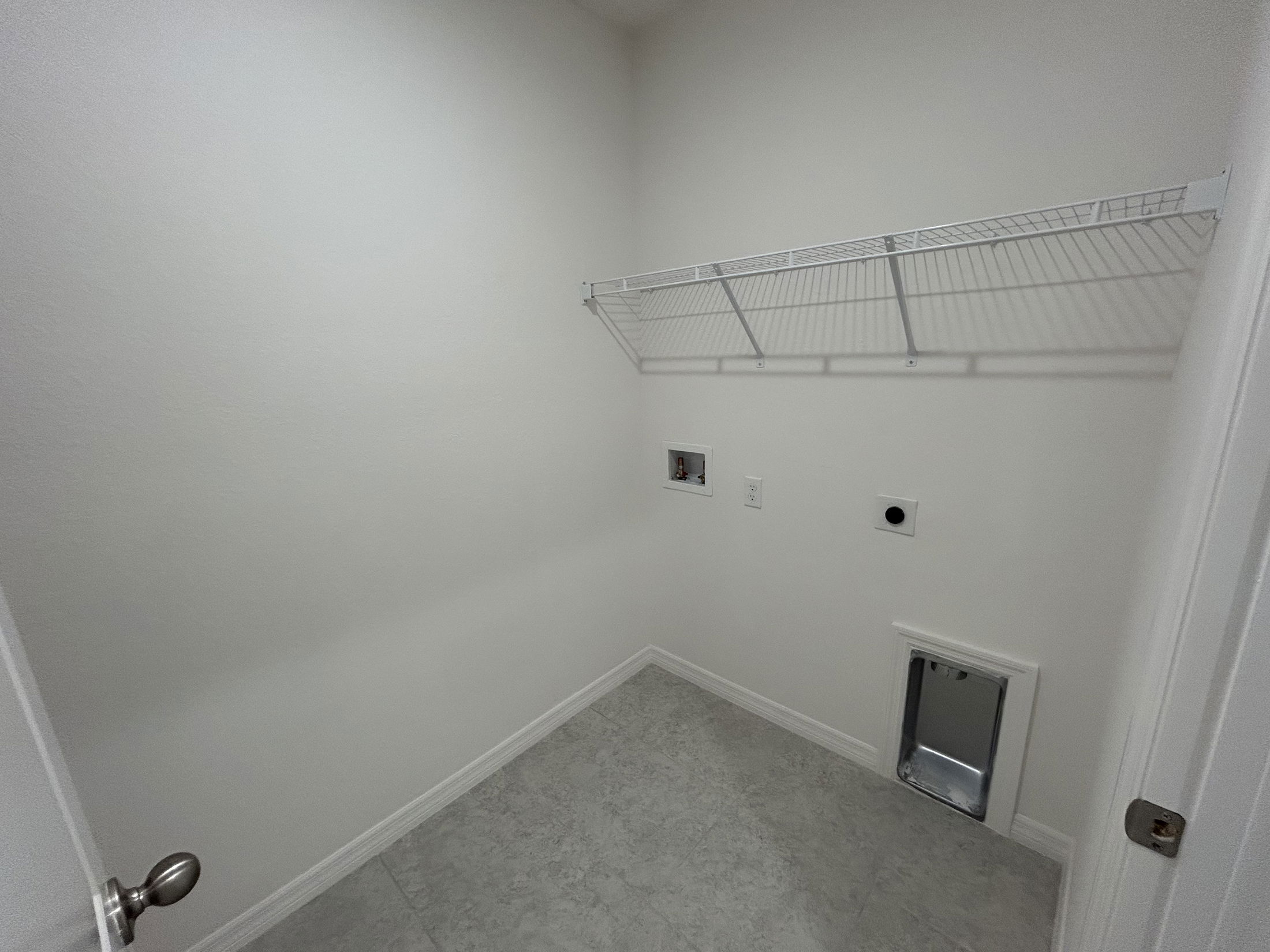 A clean and minimalist laundry room featuring a wire shelf and utility connections on white walls.
