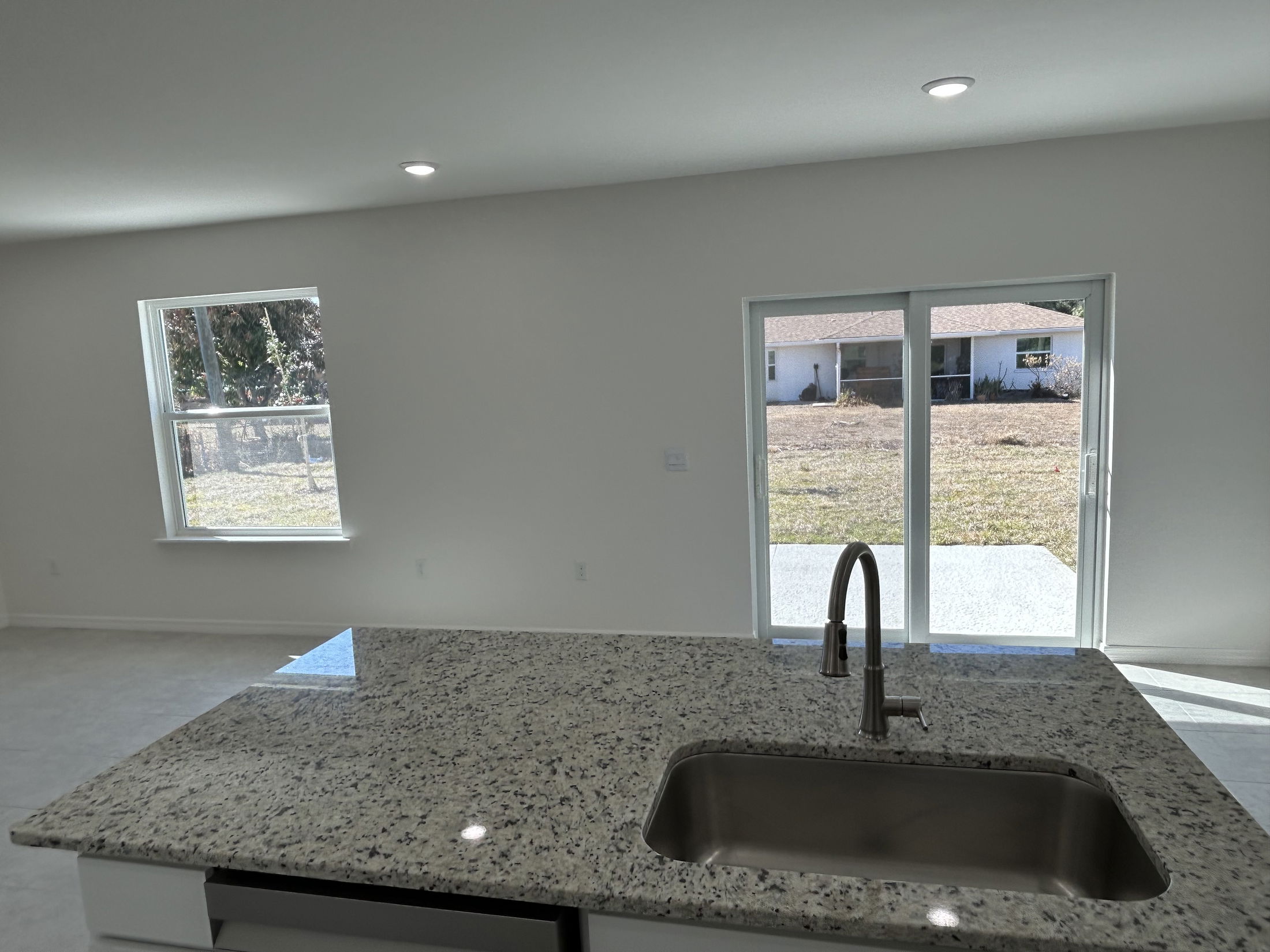 Modern kitchen interior with granite countertops, stainless steel sink, and large windows overlooking a backyard.