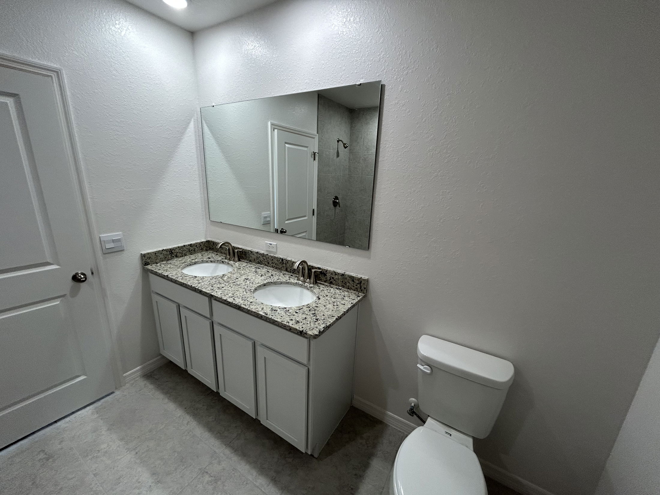Modern bathroom featuring a double sink vanity with granite countertop, large mirror, and a toilet against a neutral wall.