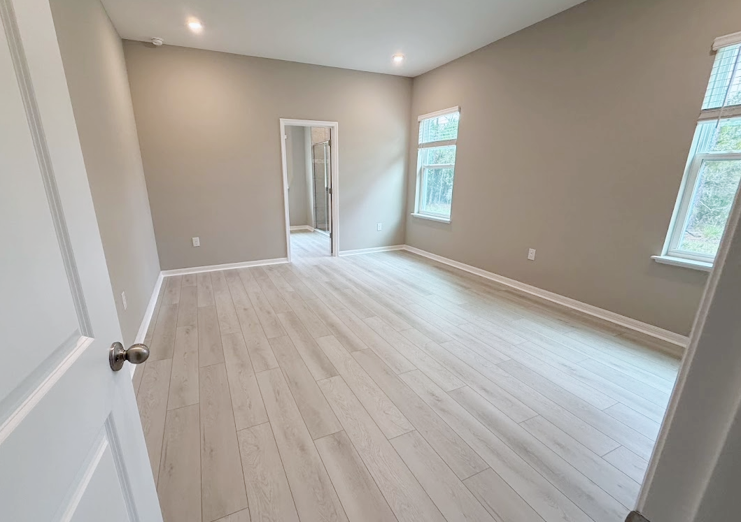 A spacious, empty room with light-colored walls and wooden flooring, featuring large windows for natural light.