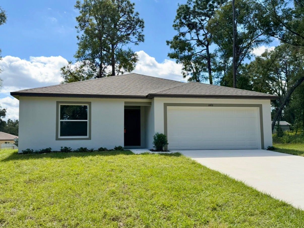 Single-story modern home with a sloped roof, two-car garage, and well-maintained lawn surrounded by trees, under a clear blue sky.