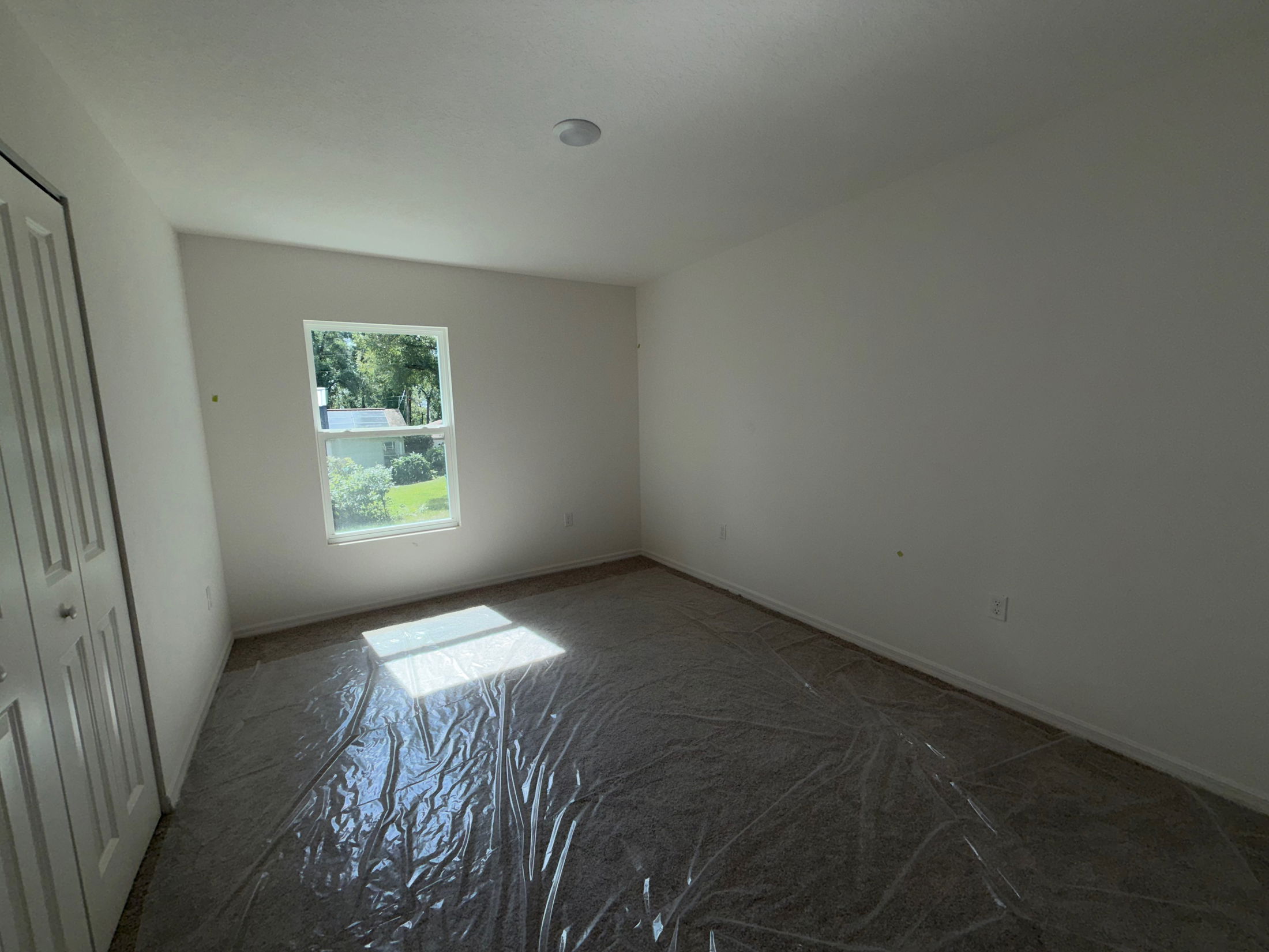 Empty room with plastic-covered carpet, white walls, a window with natural light, and a closed closet door.