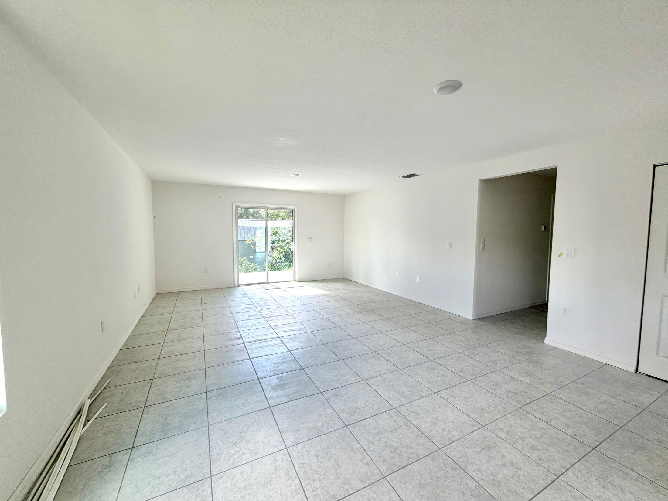 Spacious empty room with tiled floor and glass sliding door leading to a backyard garden.
