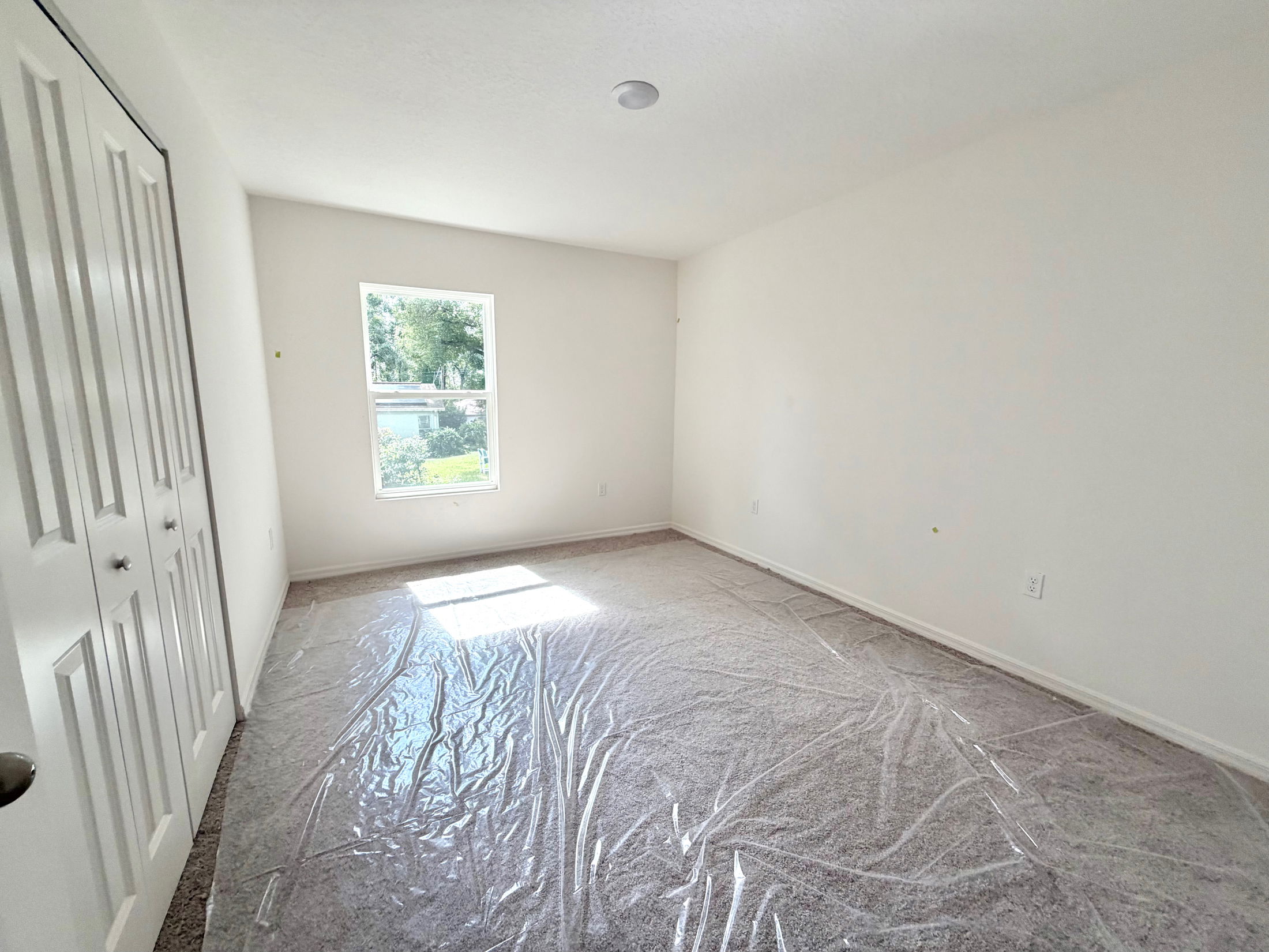 Bright, empty bedroom with new carpet and a single window, featuring closet doors and white walls.