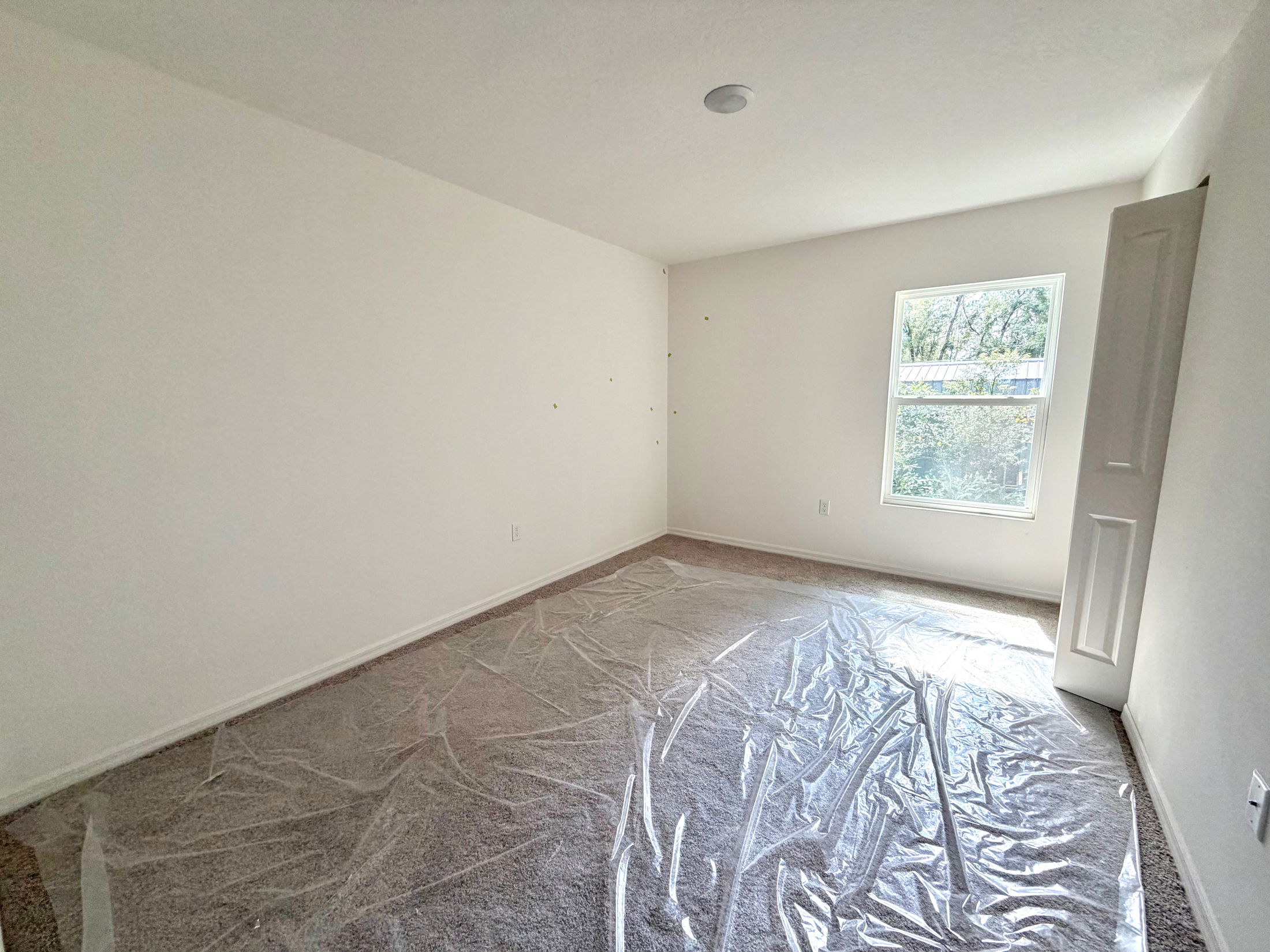 Empty white-walled room with plastic covering on the carpet and a window letting in natural light.