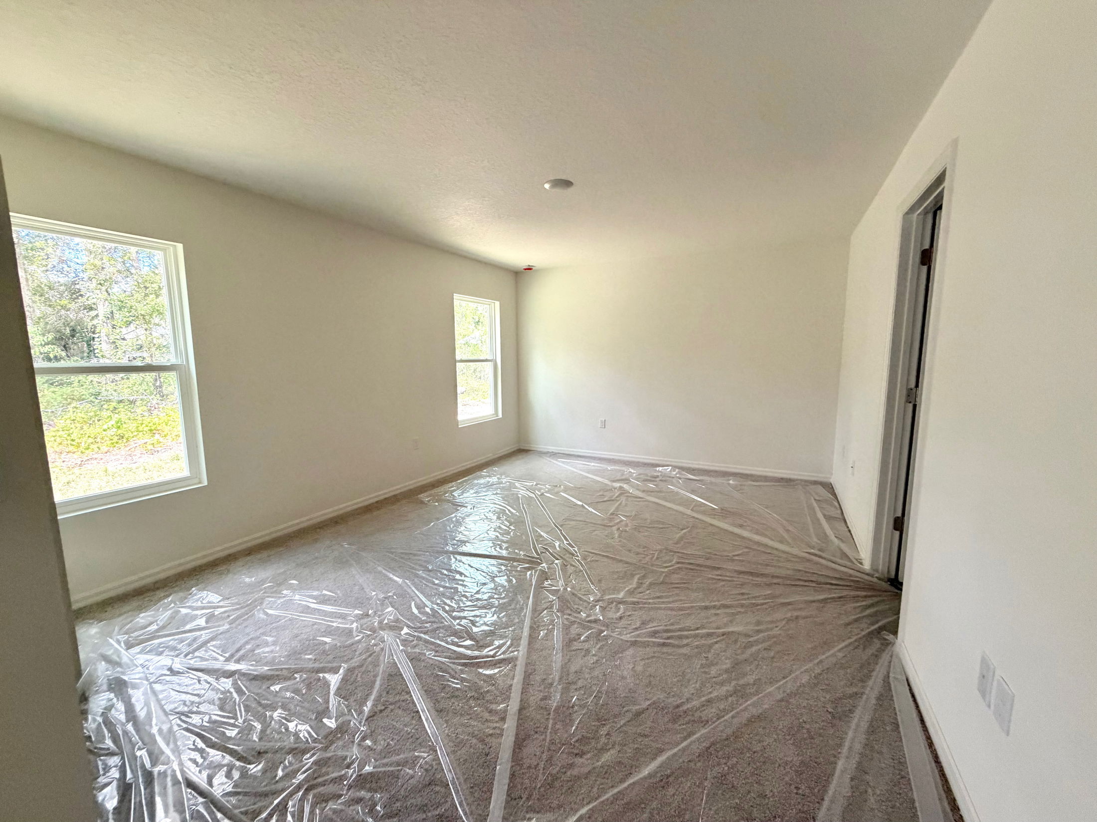 Bright, empty room with large windows, white walls, and a carpet covered in protective plastic.