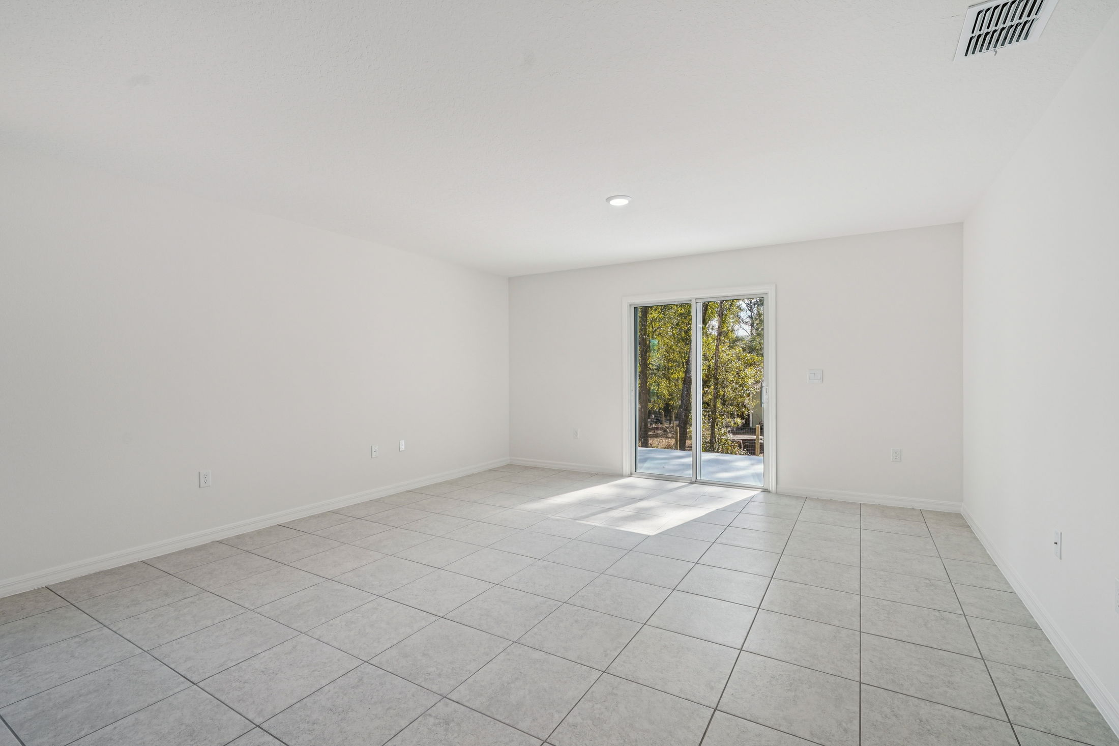 Bright and spacious living area with tile flooring and sliding glass doors leading to an outdoor deck surrounded by trees.