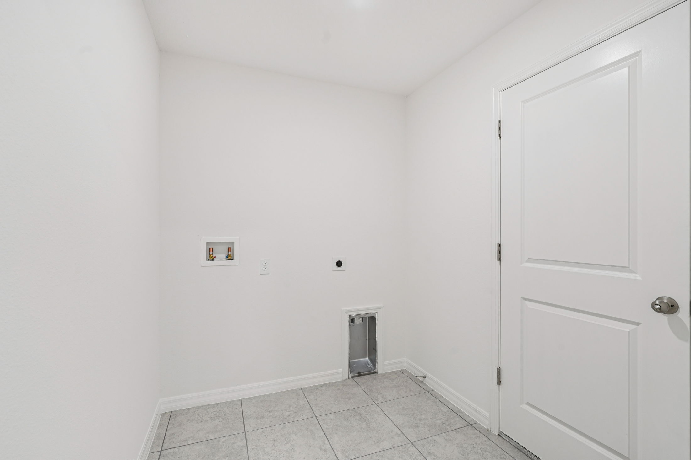 Empty laundry room with gray tile flooring and white walls, featuring plumbing connections and a door.
