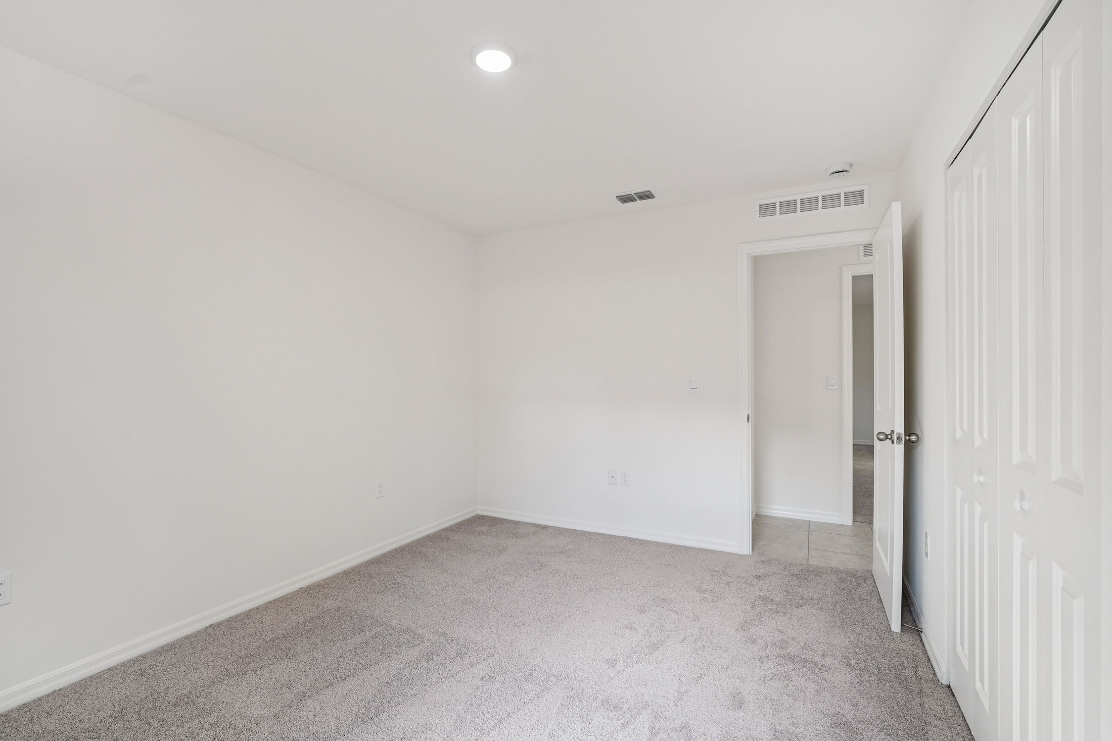 Spacious and bright empty bedroom with carpet flooring and light-colored walls, featuring a closet and open door leading to a bathroom.
