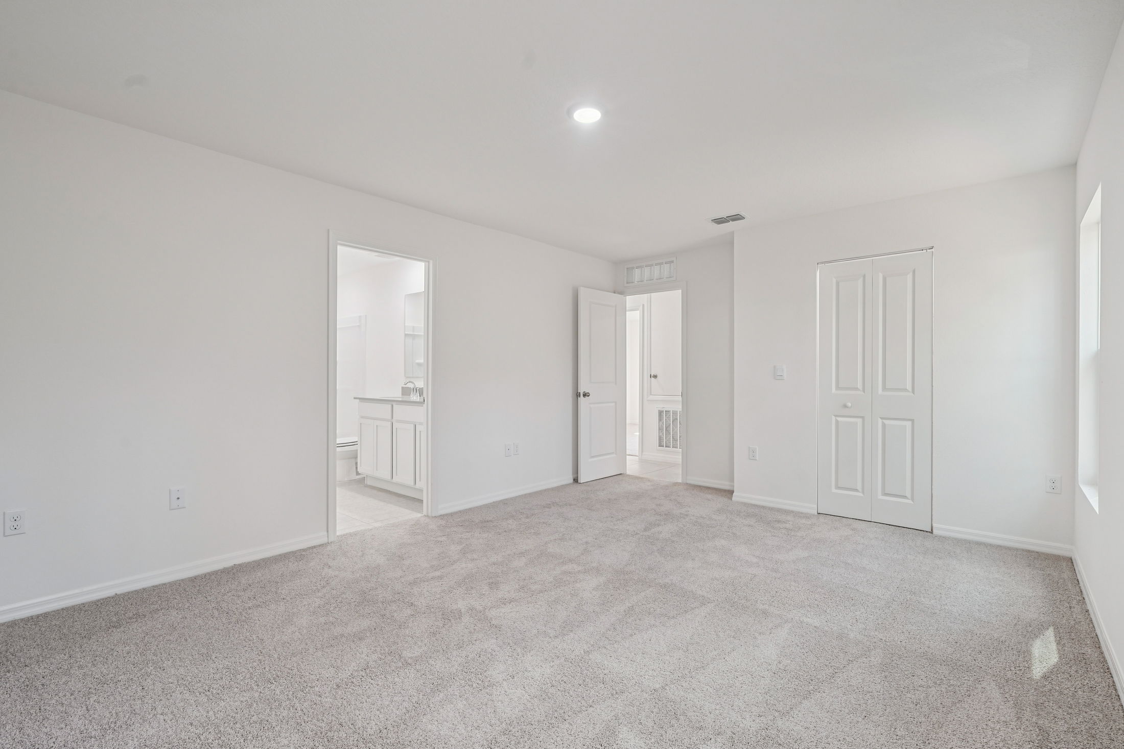 A spacious and well-lit carpeted living room featuring white walls and multiple doorways leading to adjacent rooms.