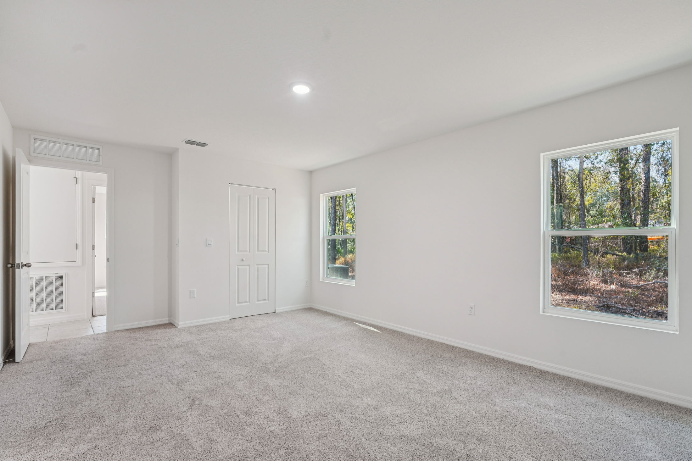 Spacious and bright empty room with soft carpet and large windows overlooking a wooded area.