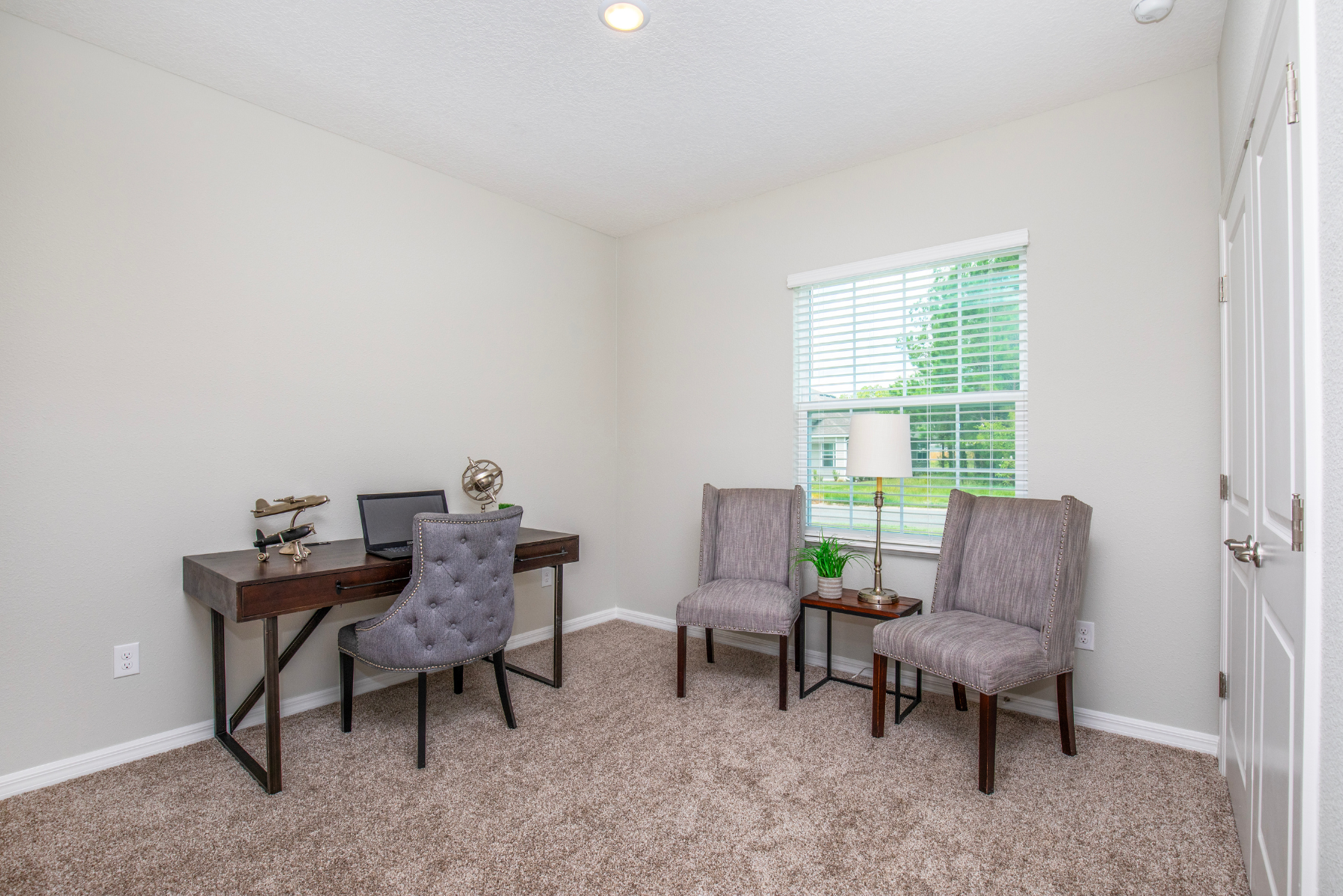 Modern home office with a brown desk, laptop, chair, two accent chairs, side table, and a window with blinds.