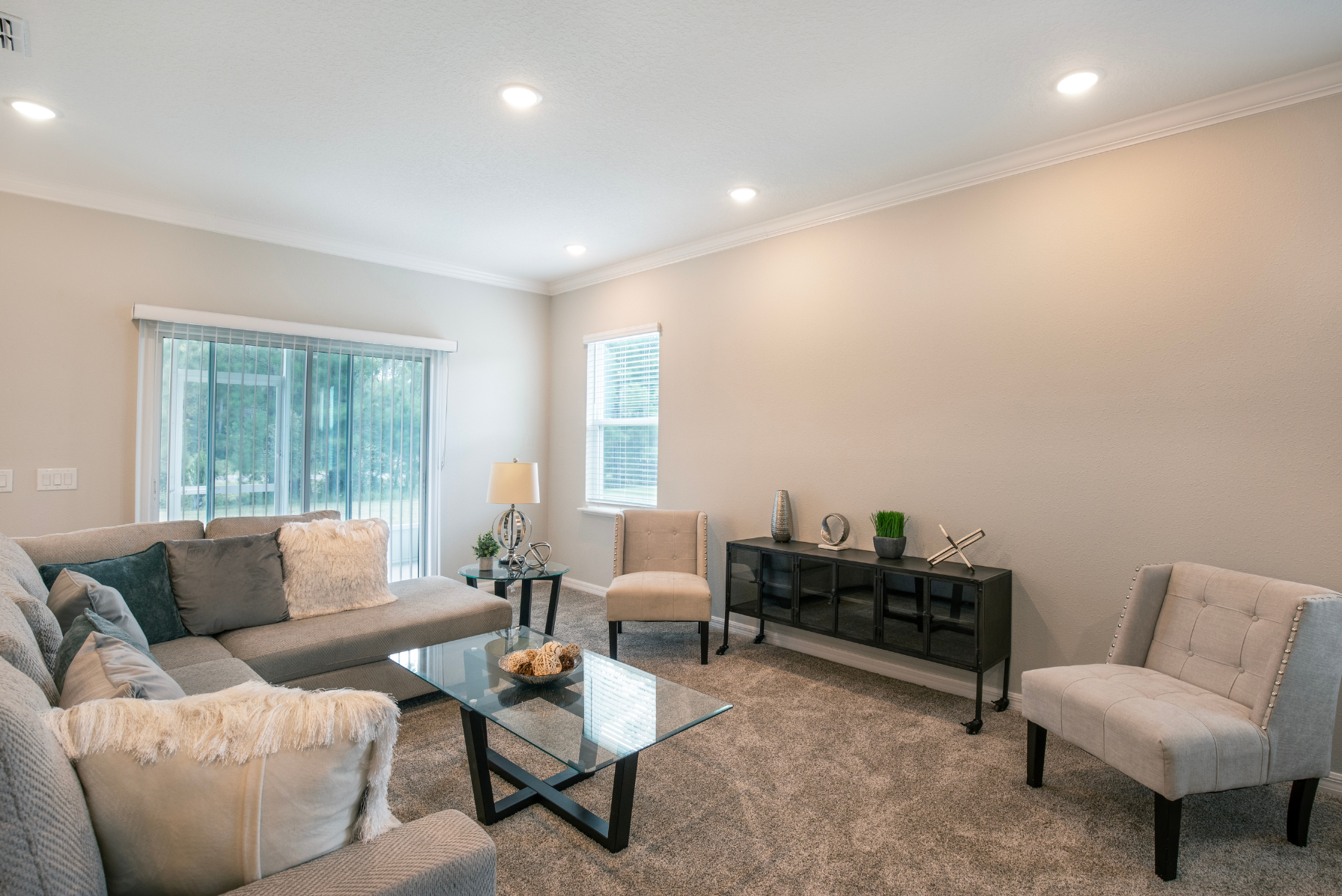 Modern living room with a gray sectional sofa, glass coffee table, and decorative accents in a bright, carpeted space.