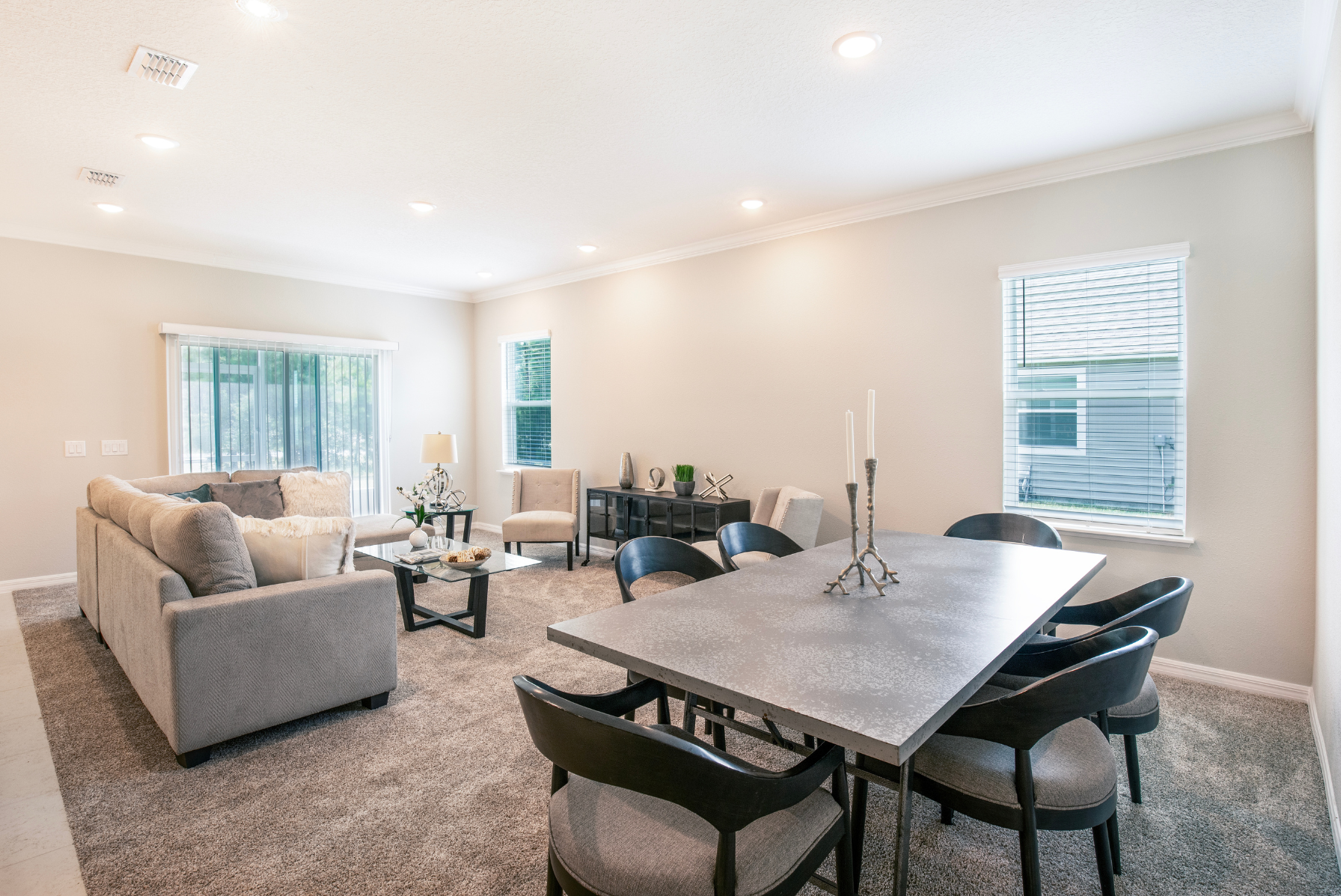 Modern open-plan living room and dining area with neutral decor, featuring a comfortable gray sofa, elegant dining table, and soft carpet flooring.