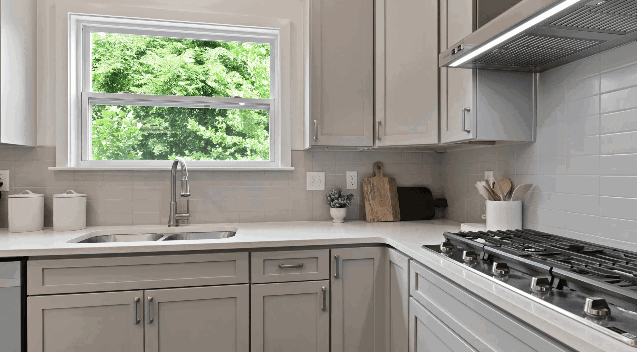 Modern kitchen interior with gray cabinets, stainless steel appliances, and a bright window overlooking greenery.