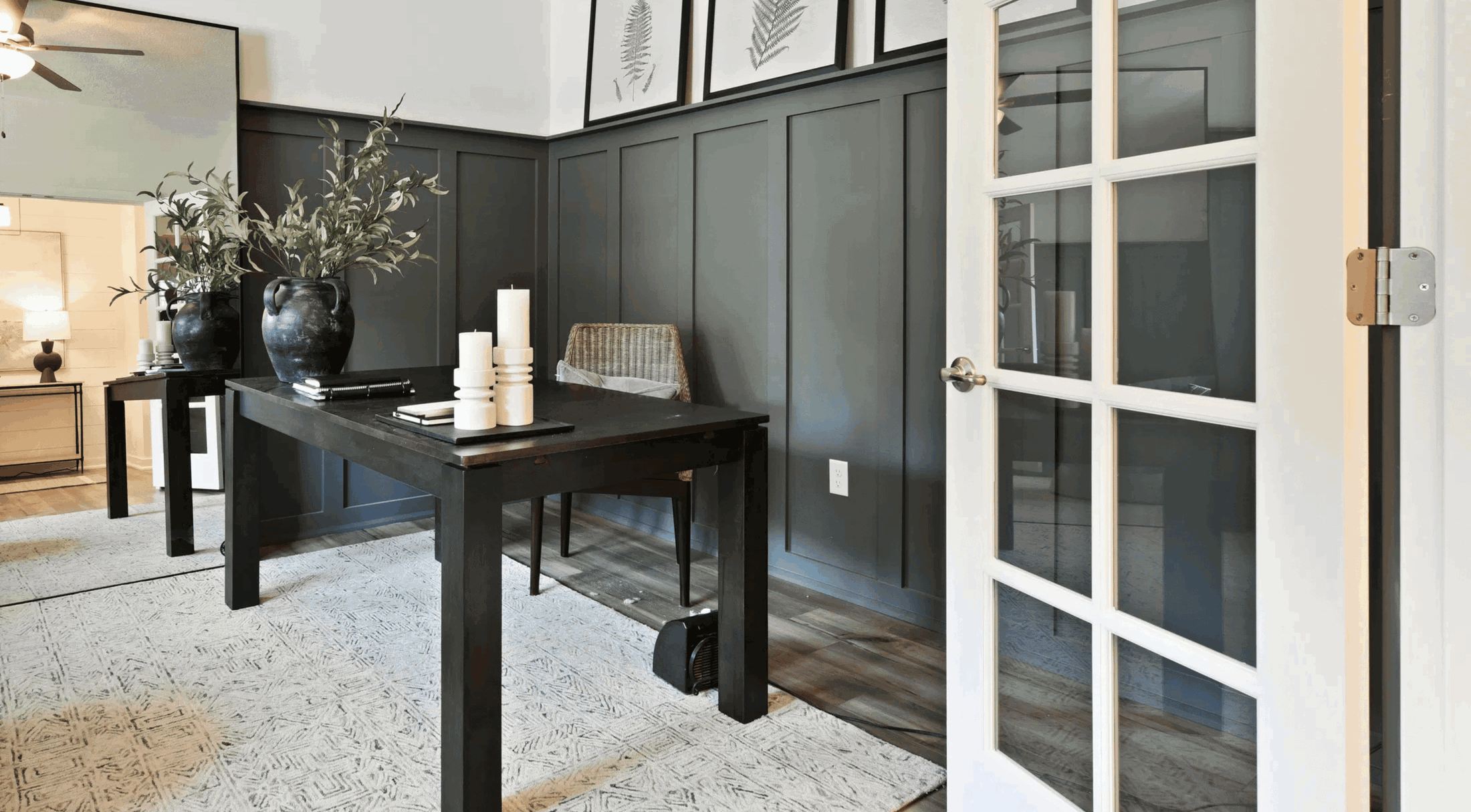 Modern home office interior with dark wood table, decorative plants, and framed artwork on gray paneled walls.