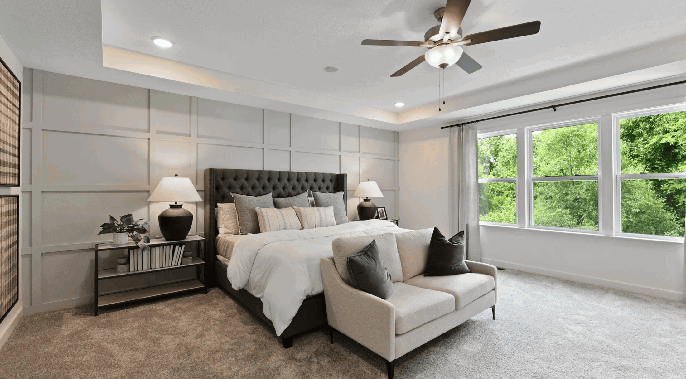 Modern bedroom interior with a stylish bed, cozy seating area, decorative lamps, and large windows showcasing a lush green view.