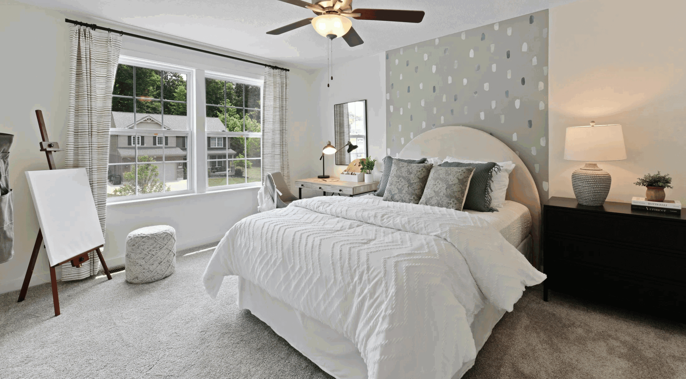 Modern bedroom featuring a stylish bed with patterned pillows, a sleek desk with a lamp by a window, and decorative accents like a pouf and an easel.