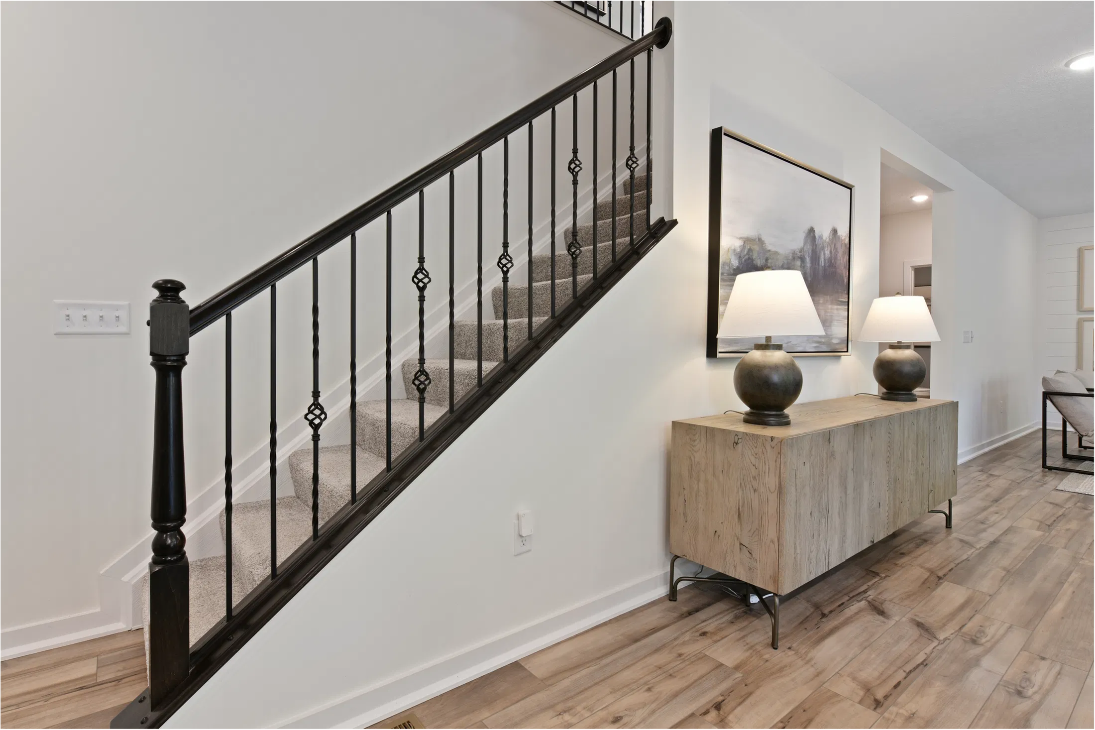 Modern interior featuring carpeted stairs with black wrought iron railing, a wooden console table with two lamps, and artwork in a bright, open hallway.