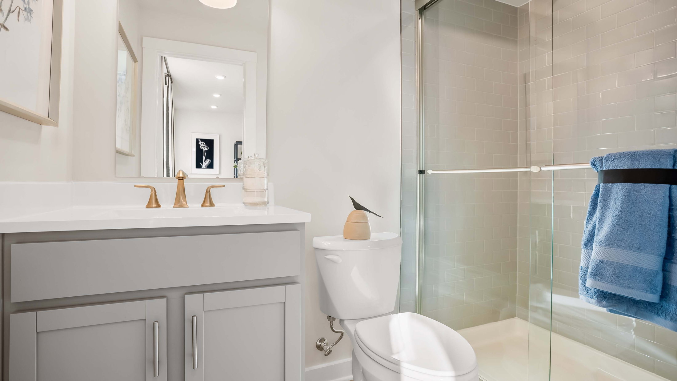 A modern bathroom featuring a gray vanity with gold faucet fixtures, a white toilet, a glass shower, and a blue towel hanging on a rod.