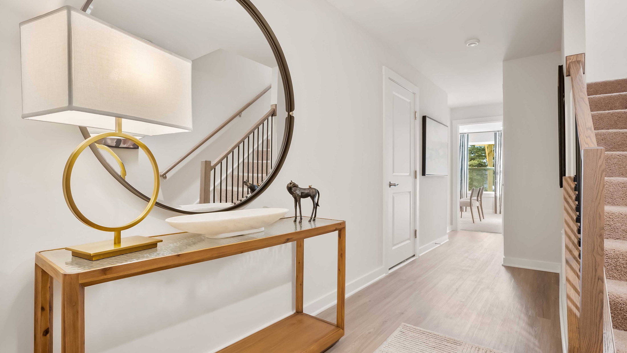A well-decorated hallway with a console table, lamp, round mirror, and staircase leading to another area.