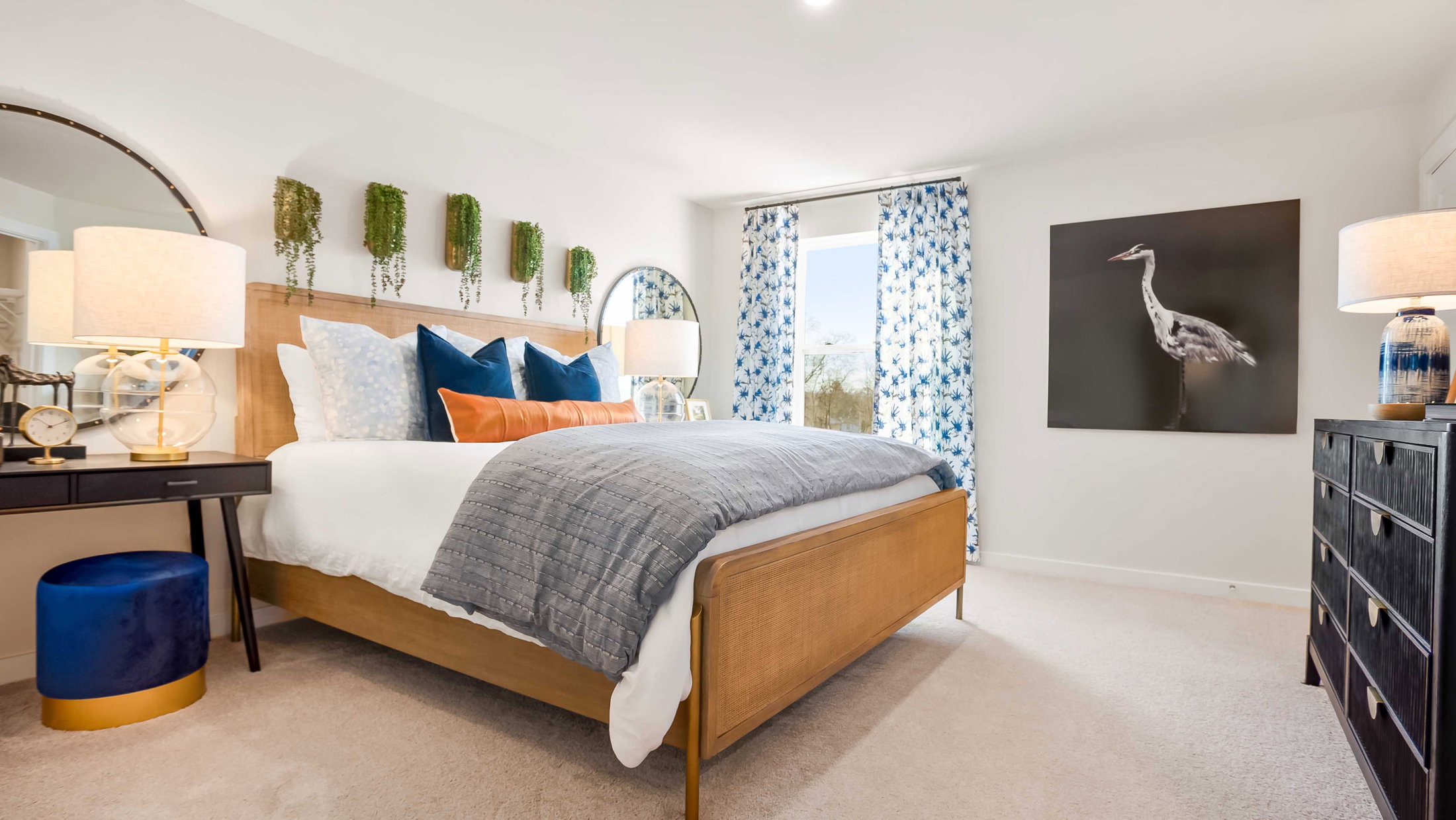 A modern bedroom featuring a wooden bed with gray and blue bedding, decorative pillows, two lamps, a circular mirror, and a large artwork of a bird on the wall.