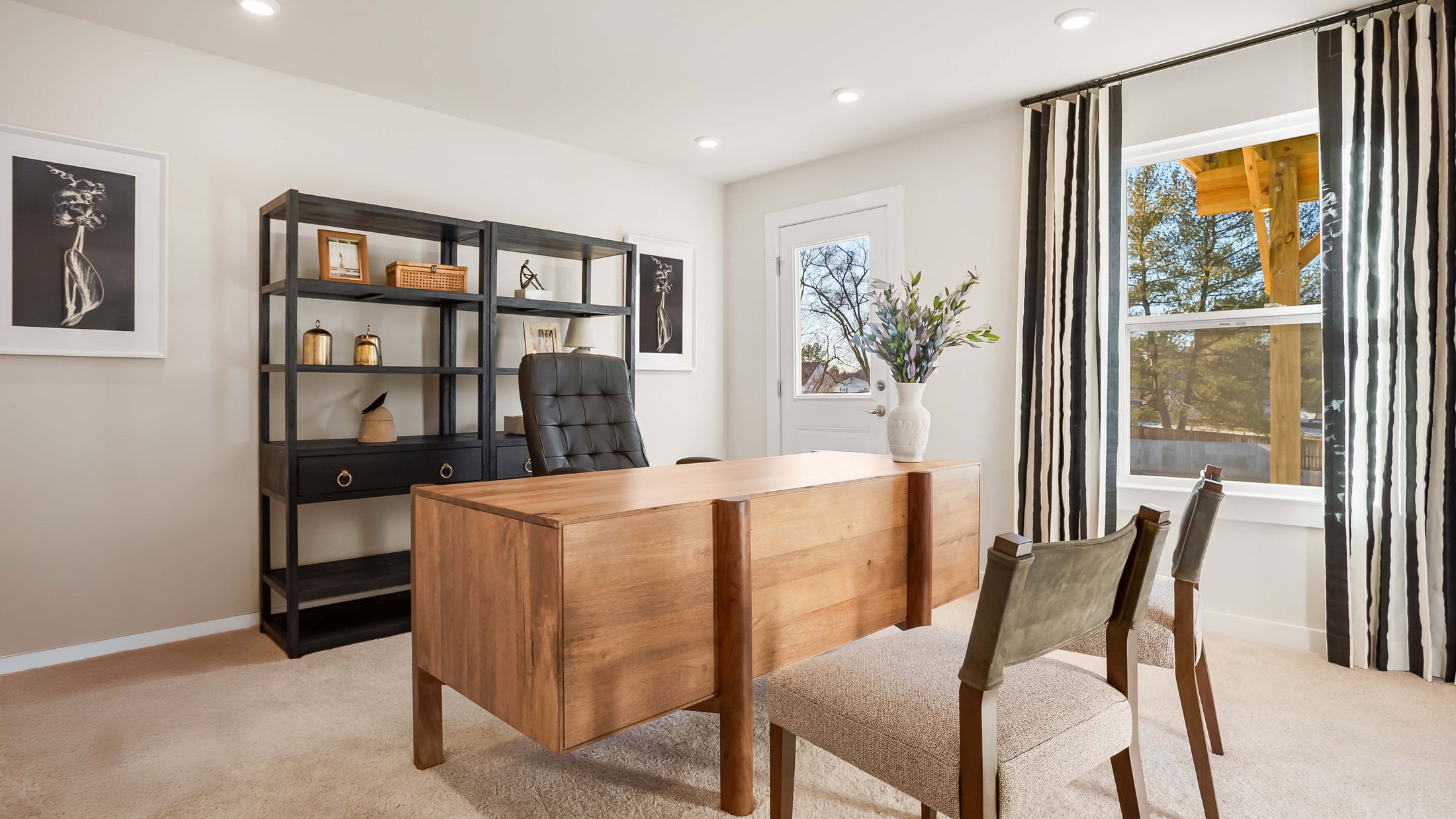 A modern office space featuring a wooden desk, black shelving unit, two chairs, and a window with striped curtains.