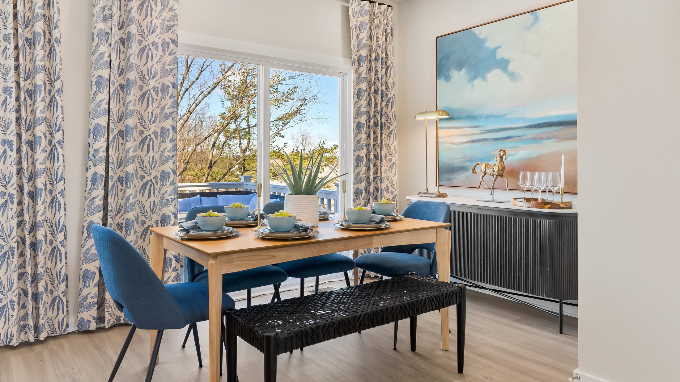A bright dining area featuring a wooden table set for four, blue chairs, a dark sideboard, and large windows with floral curtains.
