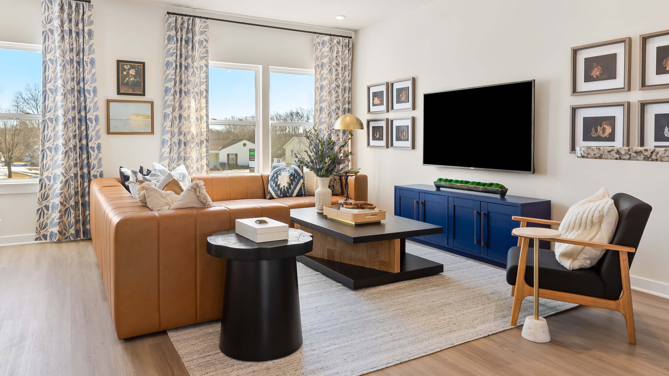 A modern living room featuring a brown leather sofa, blue sideboard, black coffee table, and large windows with light curtains.