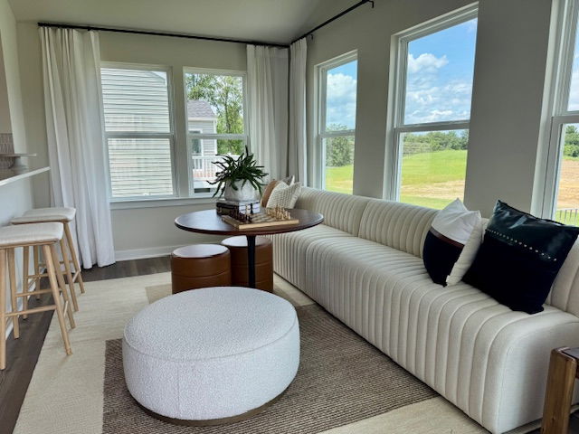 Modern sunlit living room with a white striped sofa, round ottoman, small table, and large windows overlooking a lush green landscape.