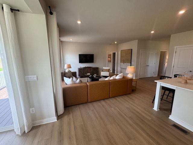 Modern open-concept living room with a brown sofa, wall-mounted TV, and wooden flooring leading to a bright kitchen area.
