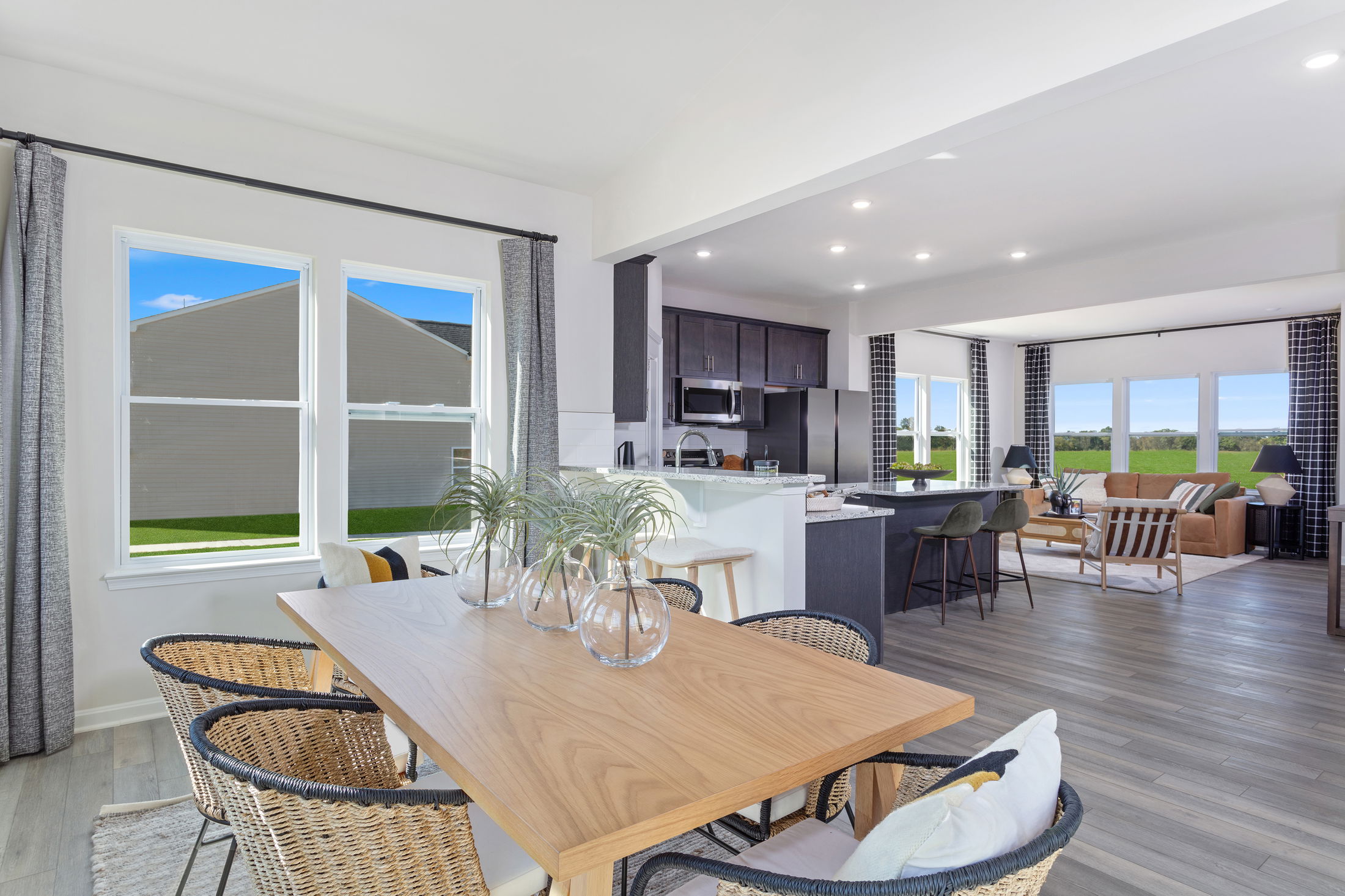 Modern open-concept living space featuring a dining area with wicker chairs, a sleek kitchen, and a bright, cozy living room with large windows.