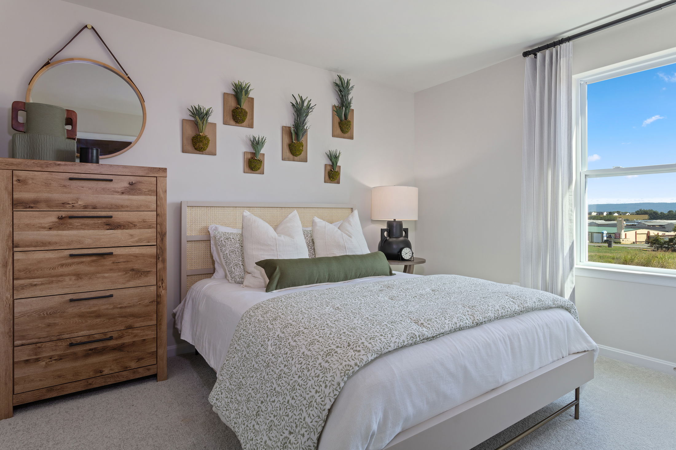 Modern bedroom with white and green bedding, a wood dresser, and a window view of a scenic landscape.