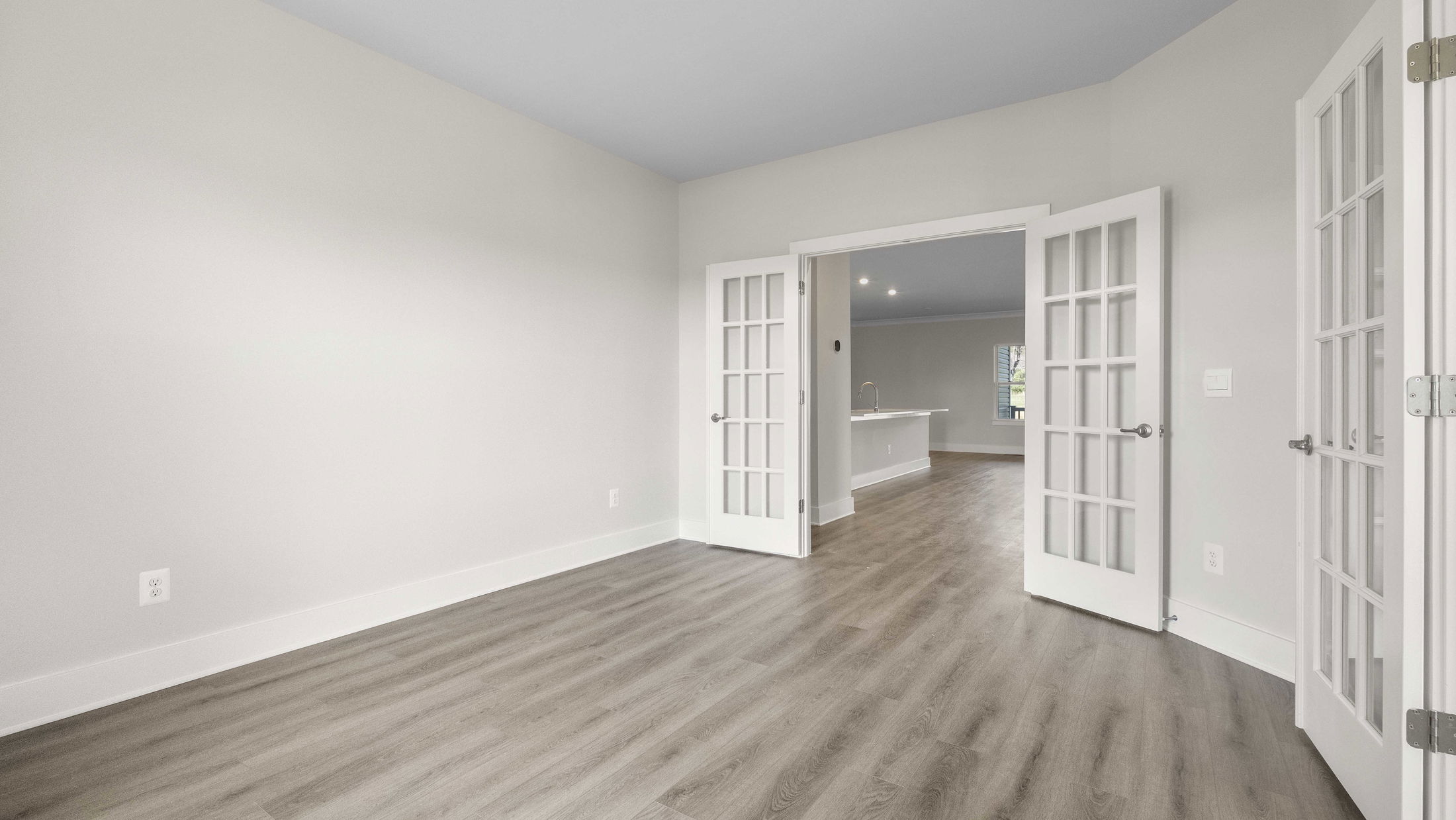 Modern, empty room with light gray walls, wood flooring, and French doors leading to a kitchen.