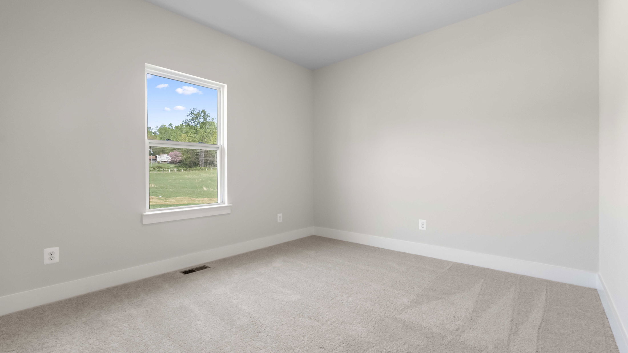 Empty room with beige walls, carpeted floor, and a window showcasing a scenic view of green fields and trees.