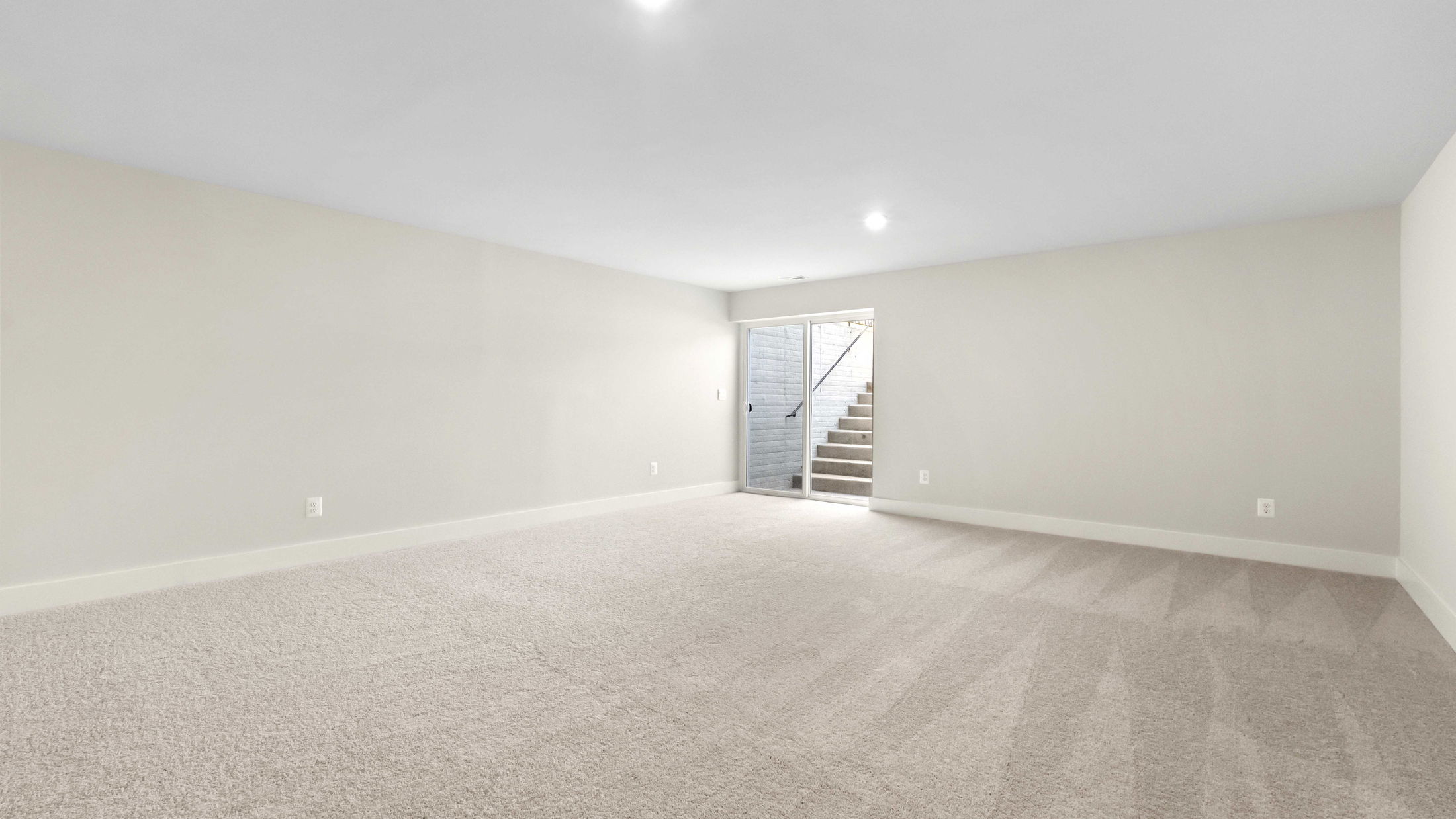 Spacious empty basement room with carpet flooring and natural light from glass doors leading to outdoor steps.