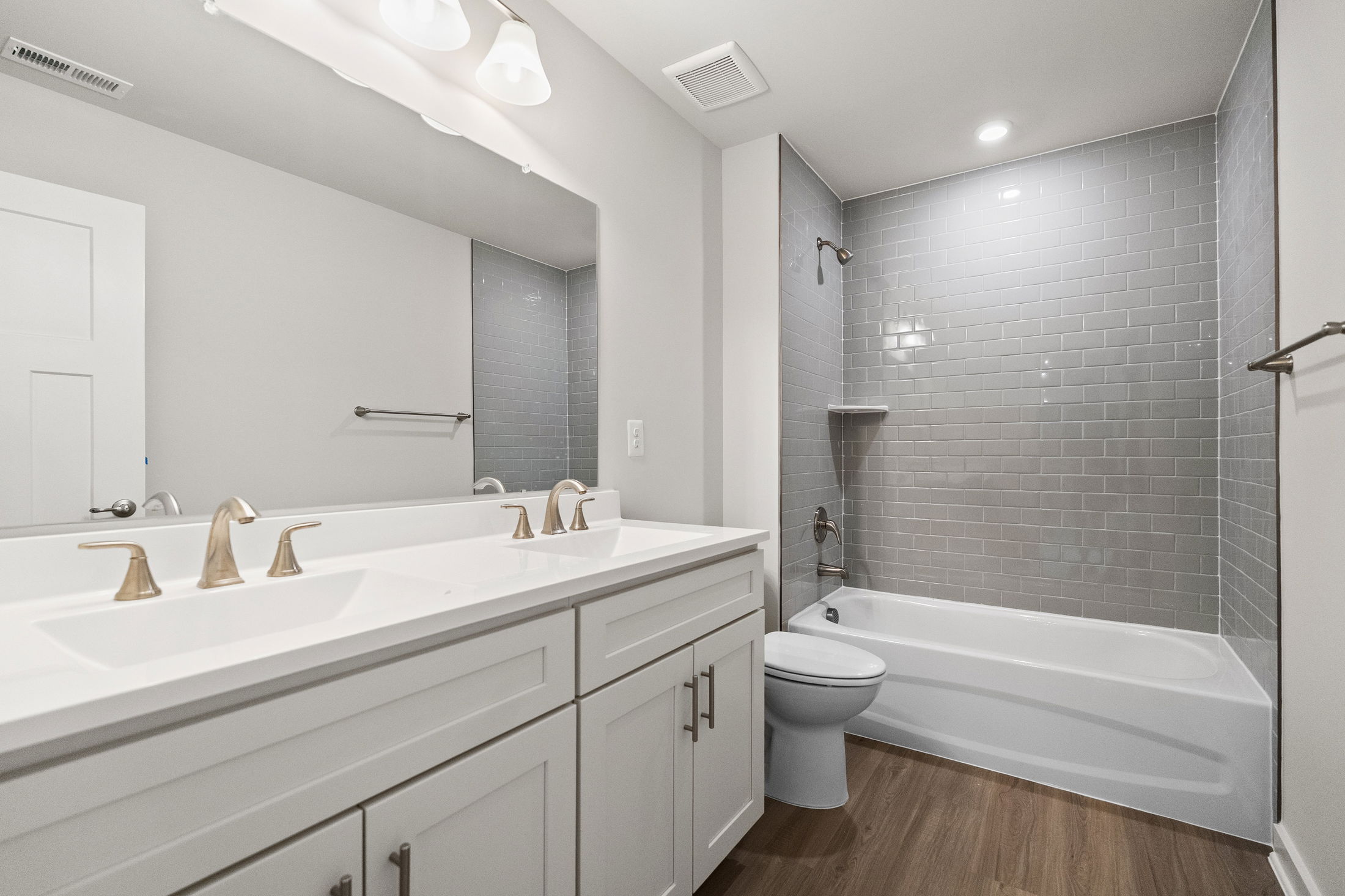 Modern bathroom featuring a double-sink vanity, light gray tile shower, and wood-style flooring.