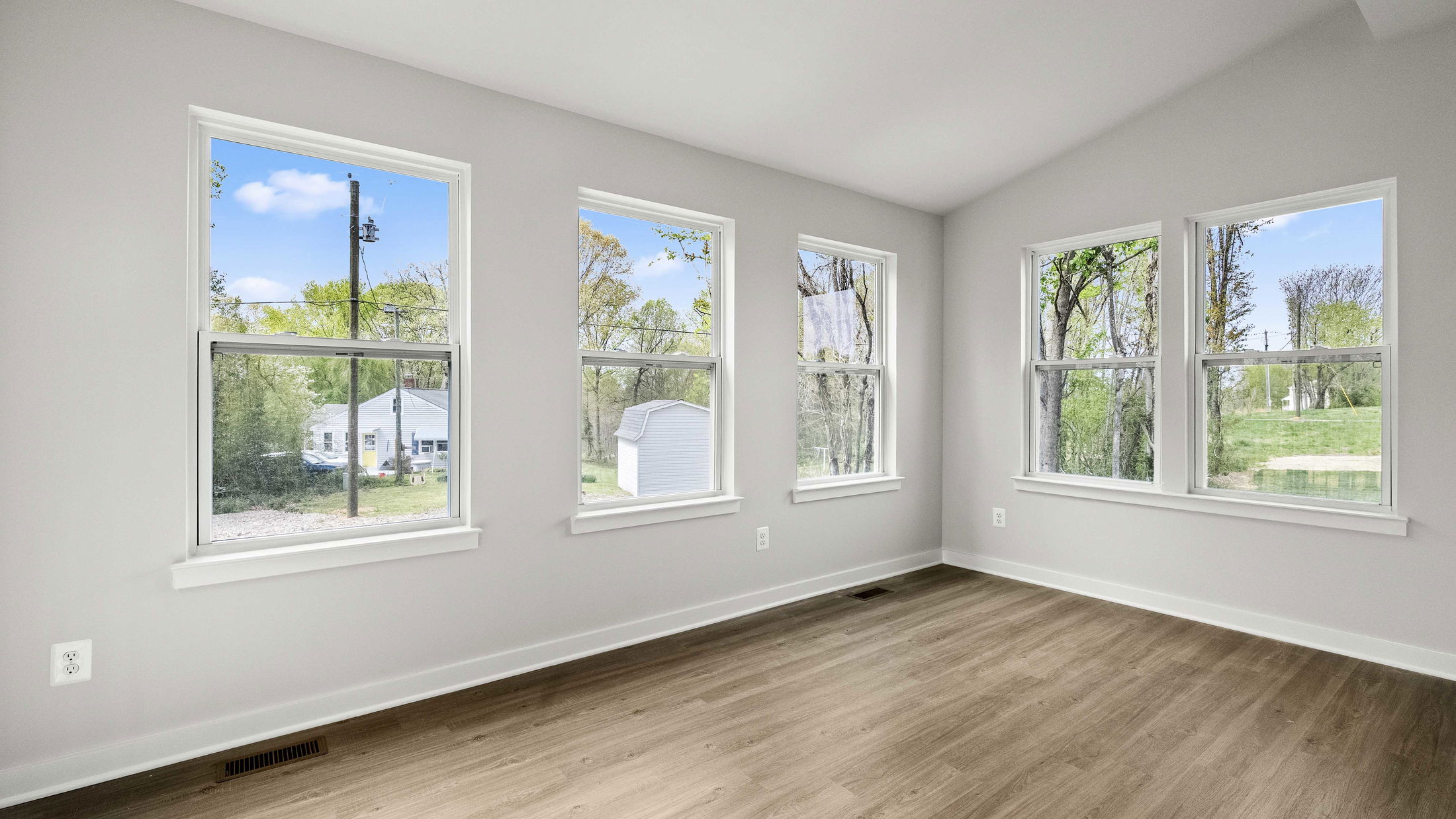 Bright room with large windows, light wood flooring, and a scenic view of trees and a white house outside.