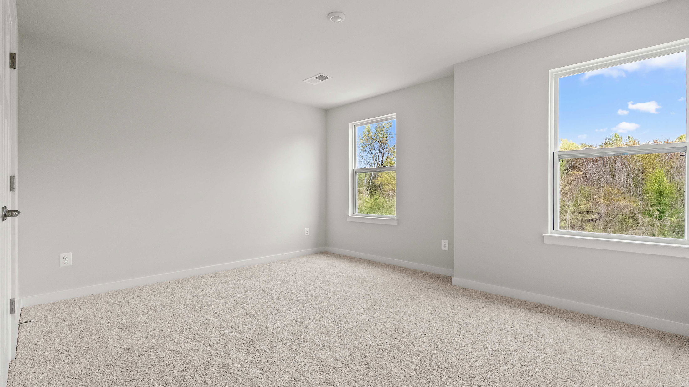 Bright, empty room with beige carpet, gray walls, and two large windows overlooking green trees and blue sky.