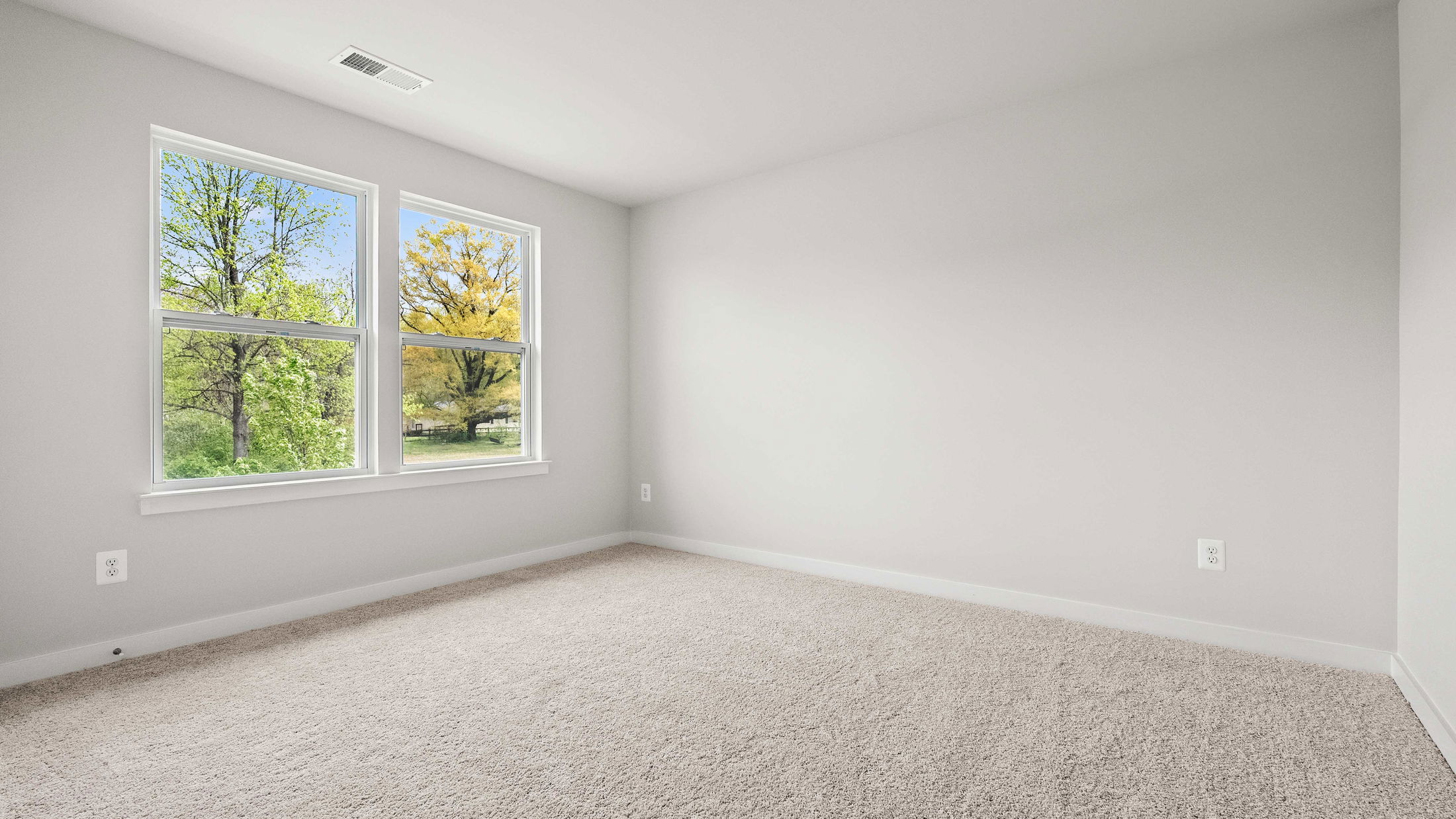Empty room with light gray walls, beige carpet, and two large windows overlooking green trees.