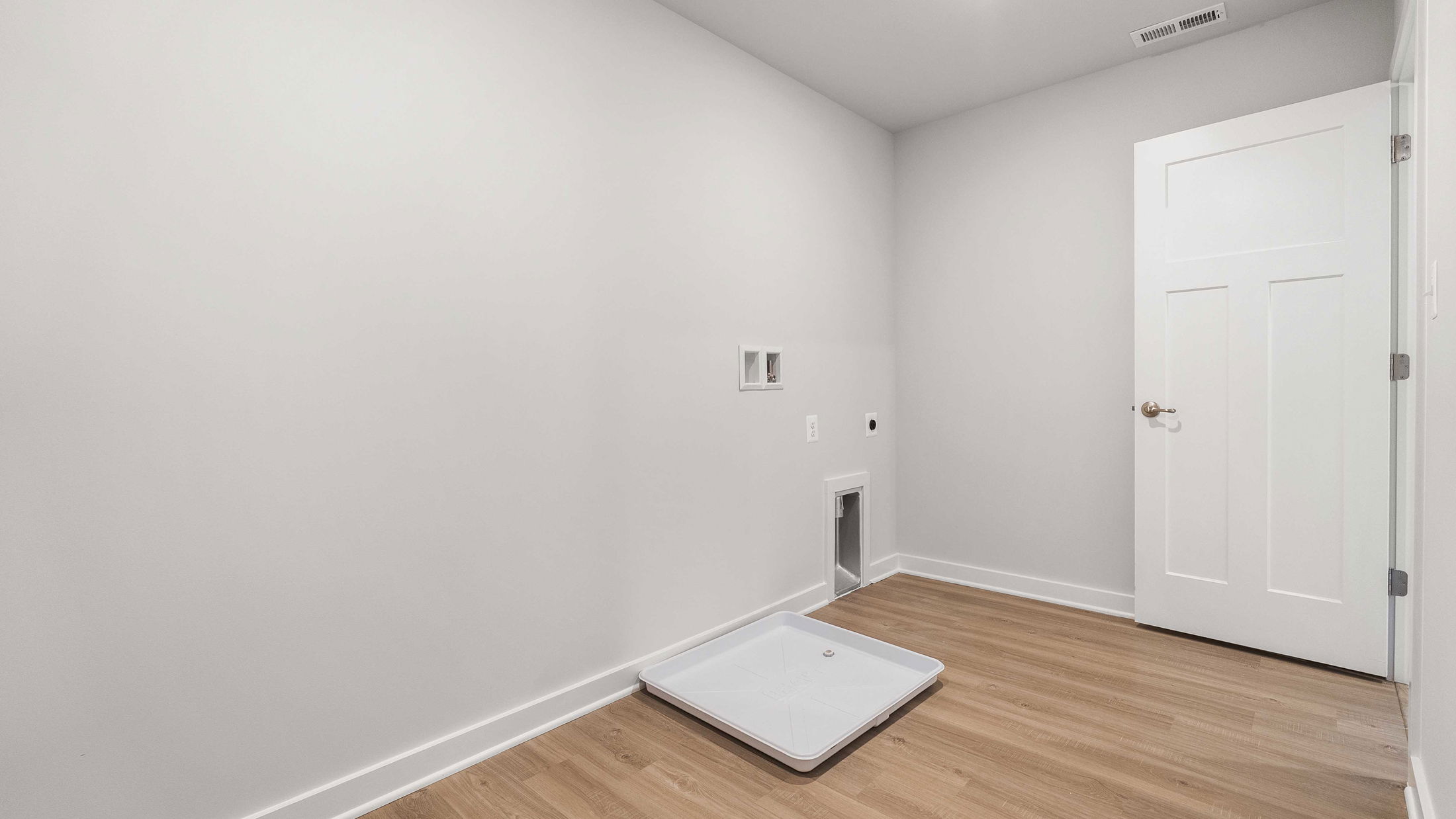 Spacious, empty laundry room with wooden floor, a white door, and utility connections on the wall.