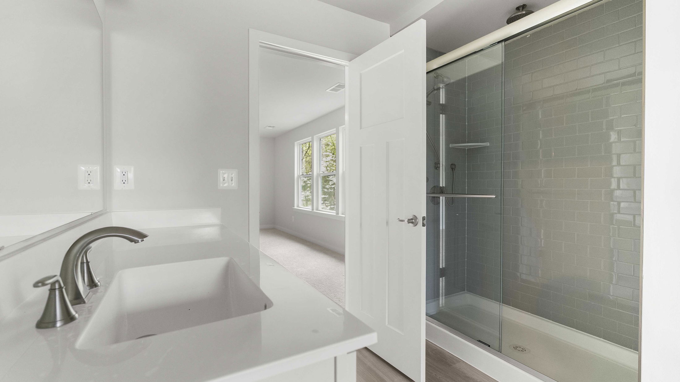 Modern bathroom with glass-enclosed shower, sleek white countertop, and chrome fixtures, leading into a well-lit bedroom.