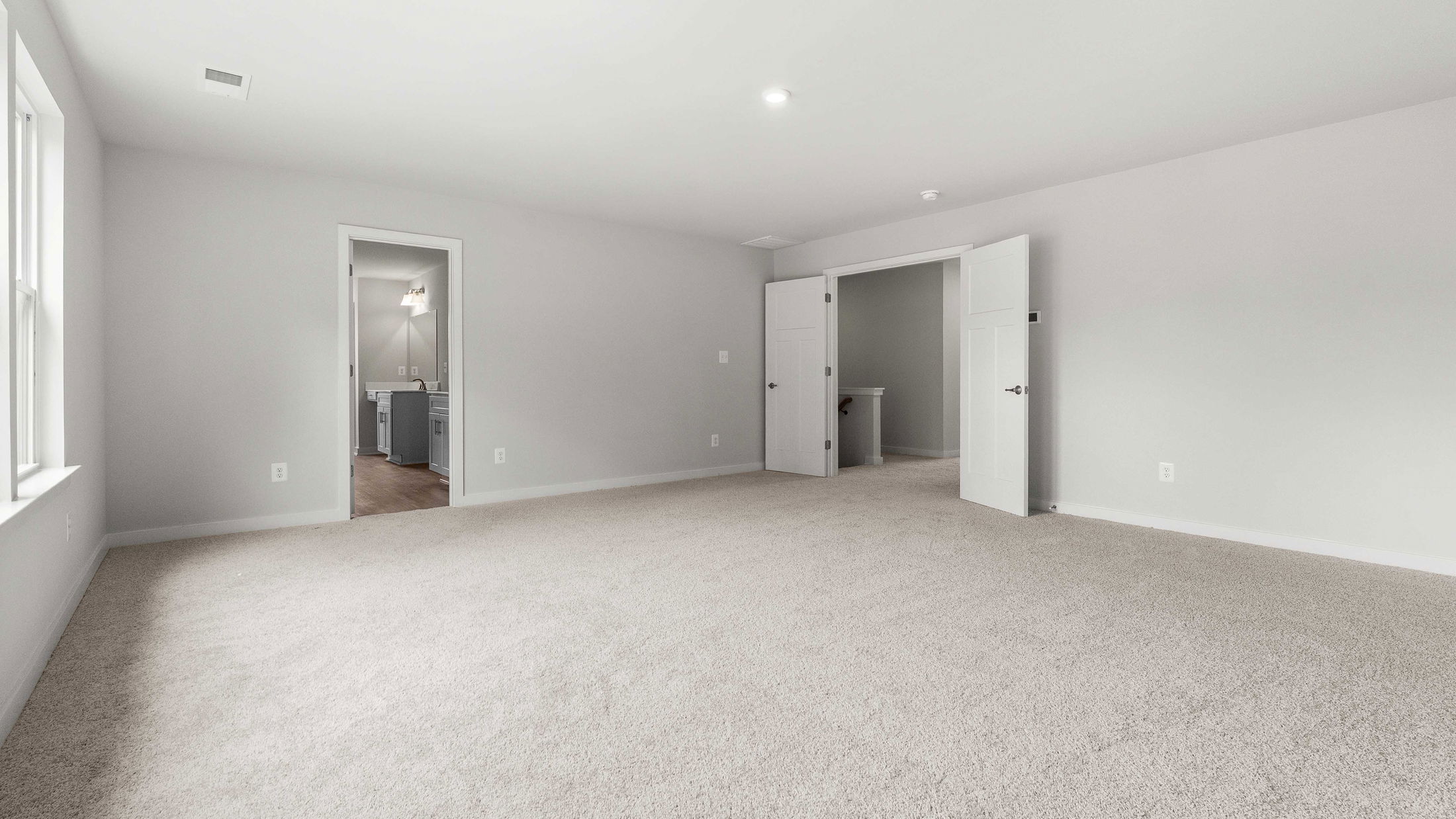 Spacious empty room with beige carpet, white walls, and an open door revealing a vanity area with hardwood floors.