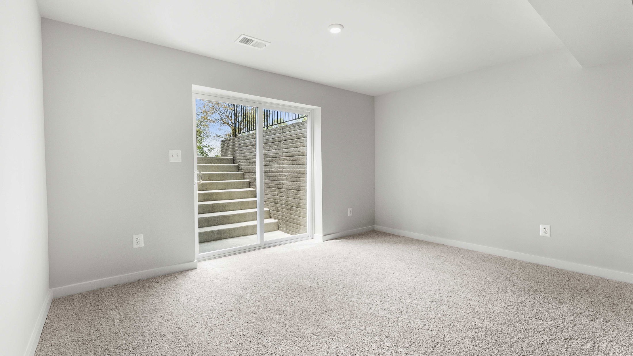 Bright, modern basement room with carpet flooring and sliding glass doors leading to outdoor stairs.