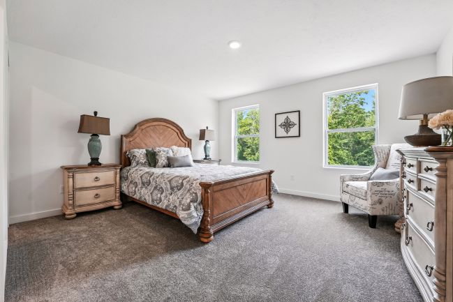 A well-lit bedroom featuring a wooden bed frame, two windows, a nightstand with a lamp, and an accent chair.