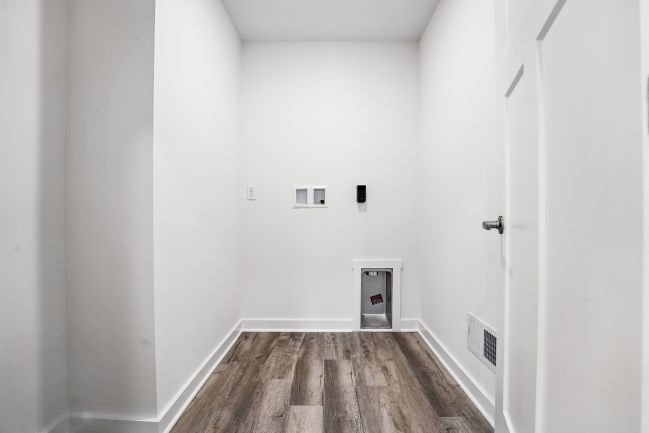 Empty white room with a door, hardwood floor, wall sockets, and a small vent.