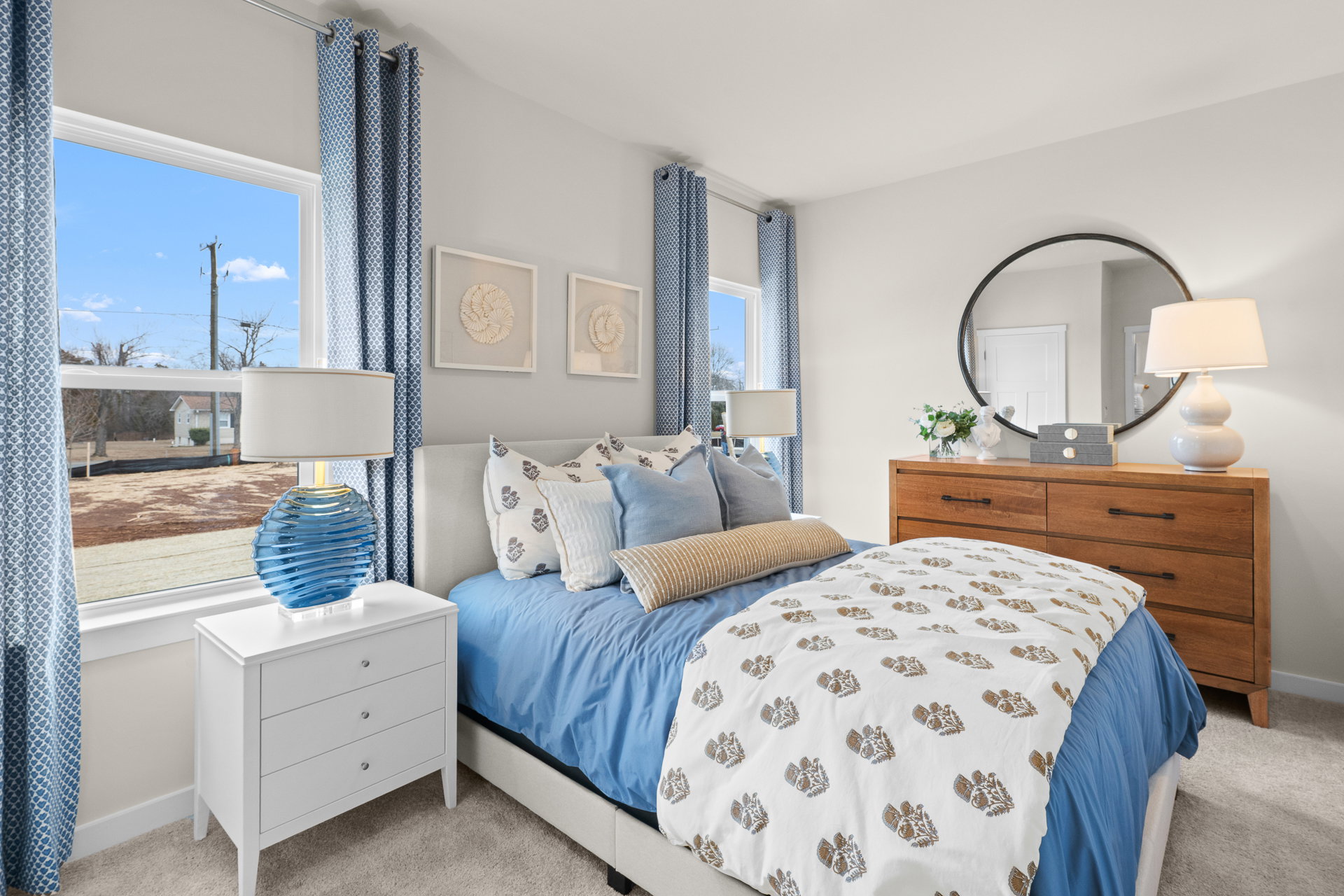 A cozy bedroom featuring a light blue comforter with decorative pillows, a wooden dresser, a round mirror, and large windows with blue curtains letting in natural light.