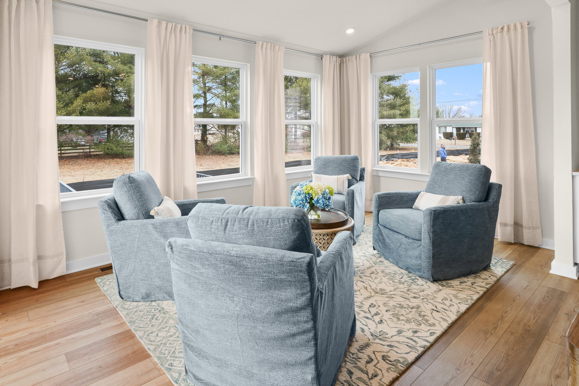 A cozy living area featuring two blue armchairs, a small table with flowers, and large windows with light curtains.